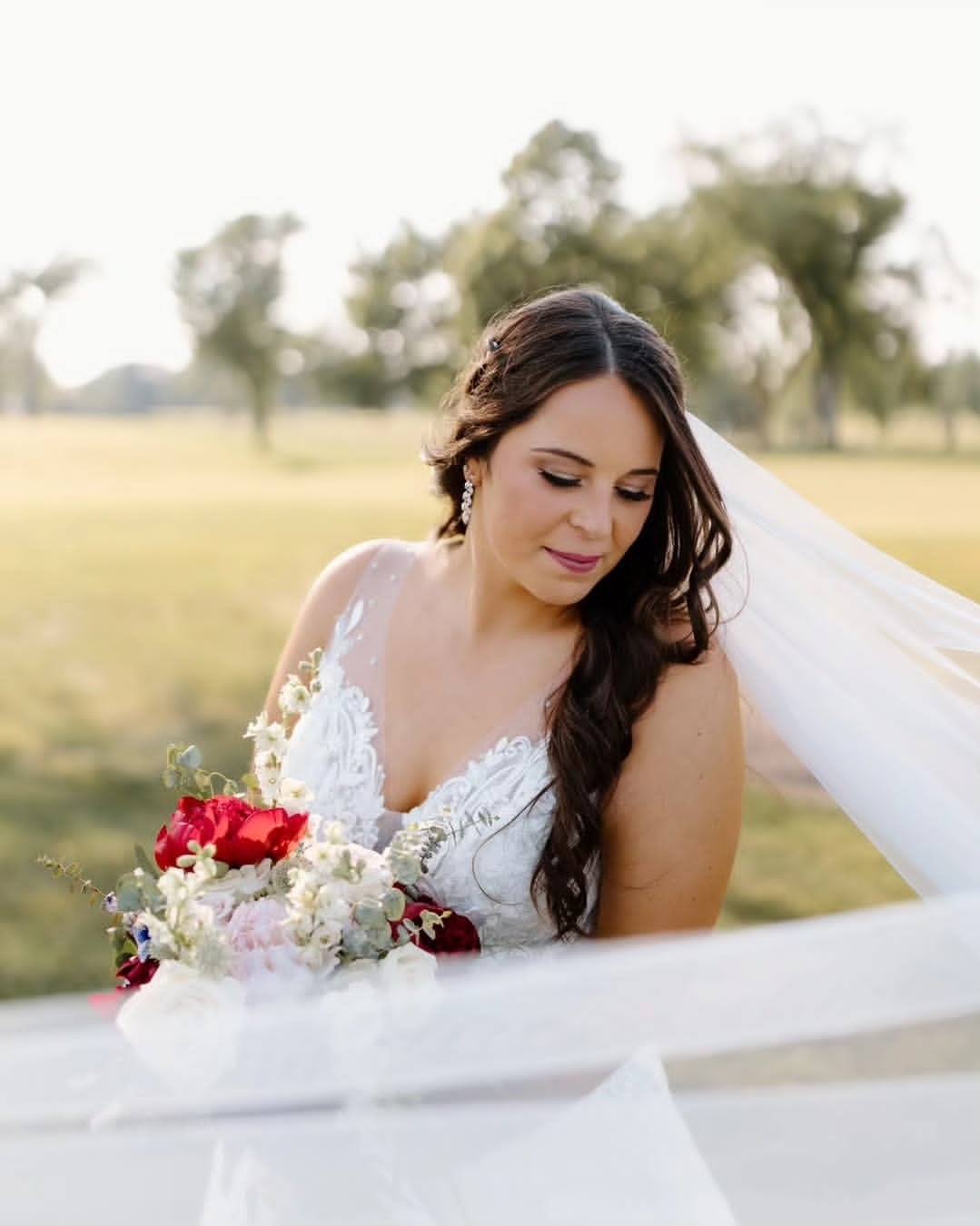 Bride with long dark hair holding a bouquet of red and white flowers outdoors, looking down, with trees in the background.