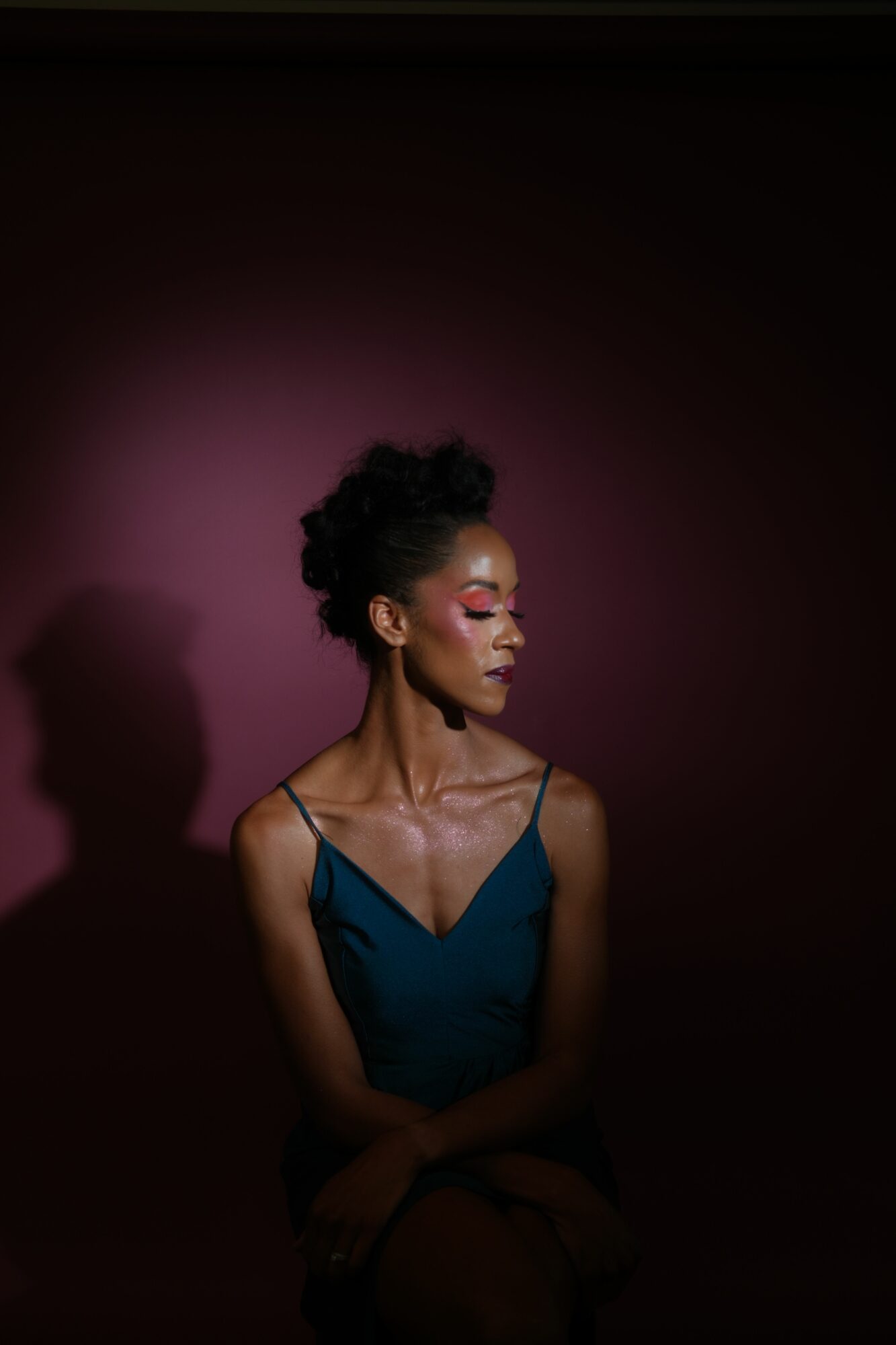 Woman with short curly hair, wearing a blue top, looking down, with pink and purple lighting, against a dark background.