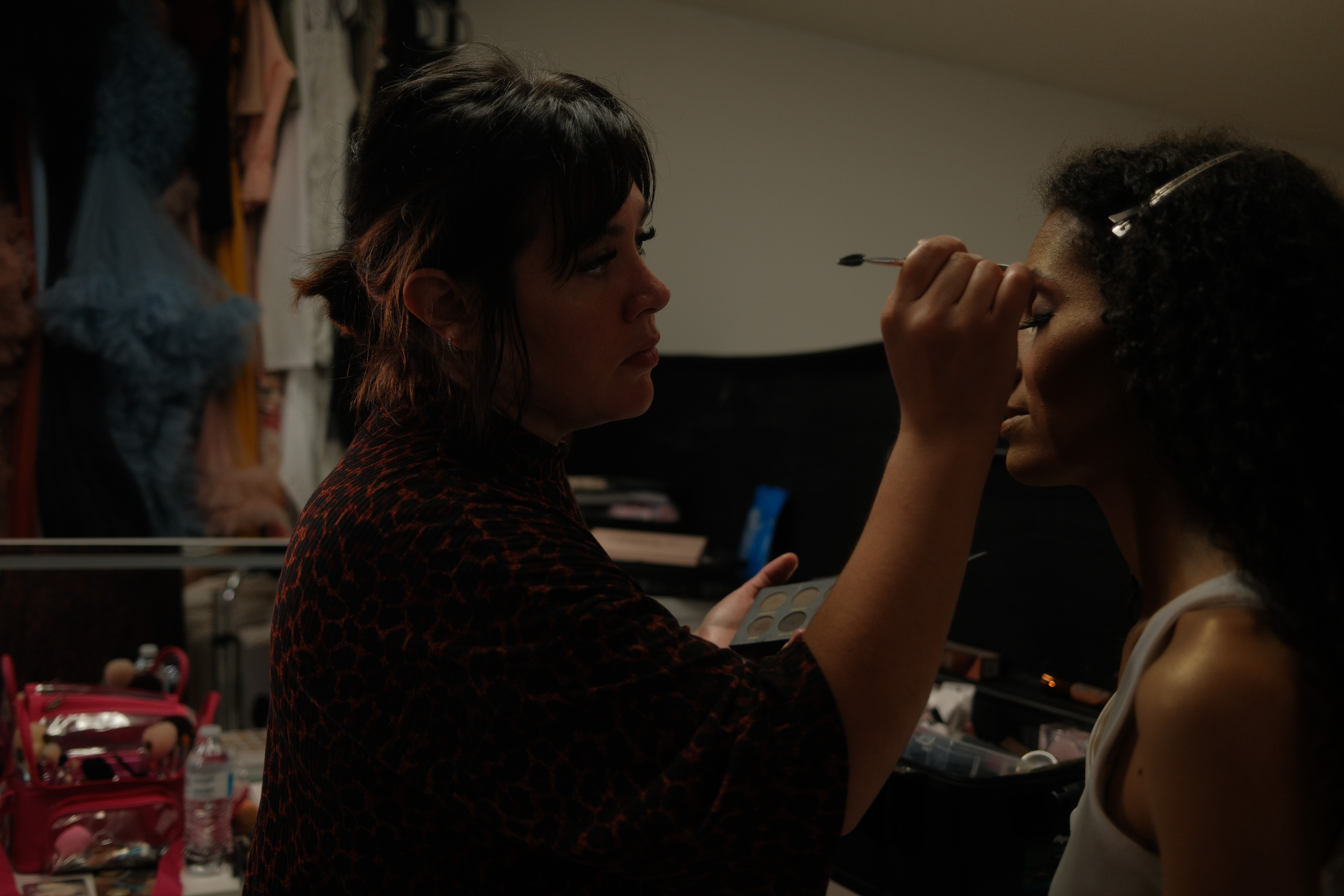 Makeup artist applying makeup to a young woman in a room with clothing and makeup items visible.
