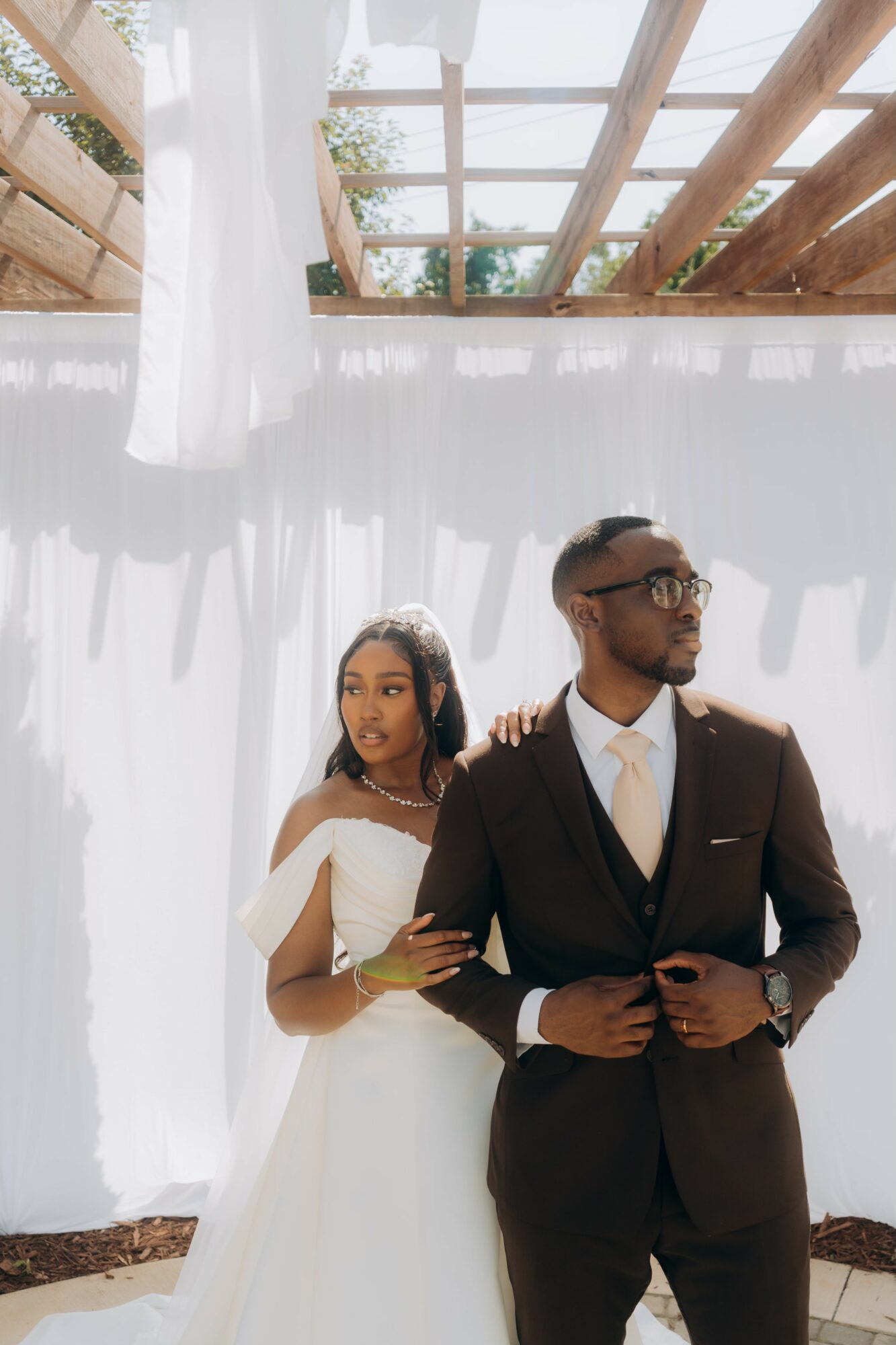 A couple dressed in wedding attire standing outdoors under a wooden pergola with white curtains, sunlight filtering through.