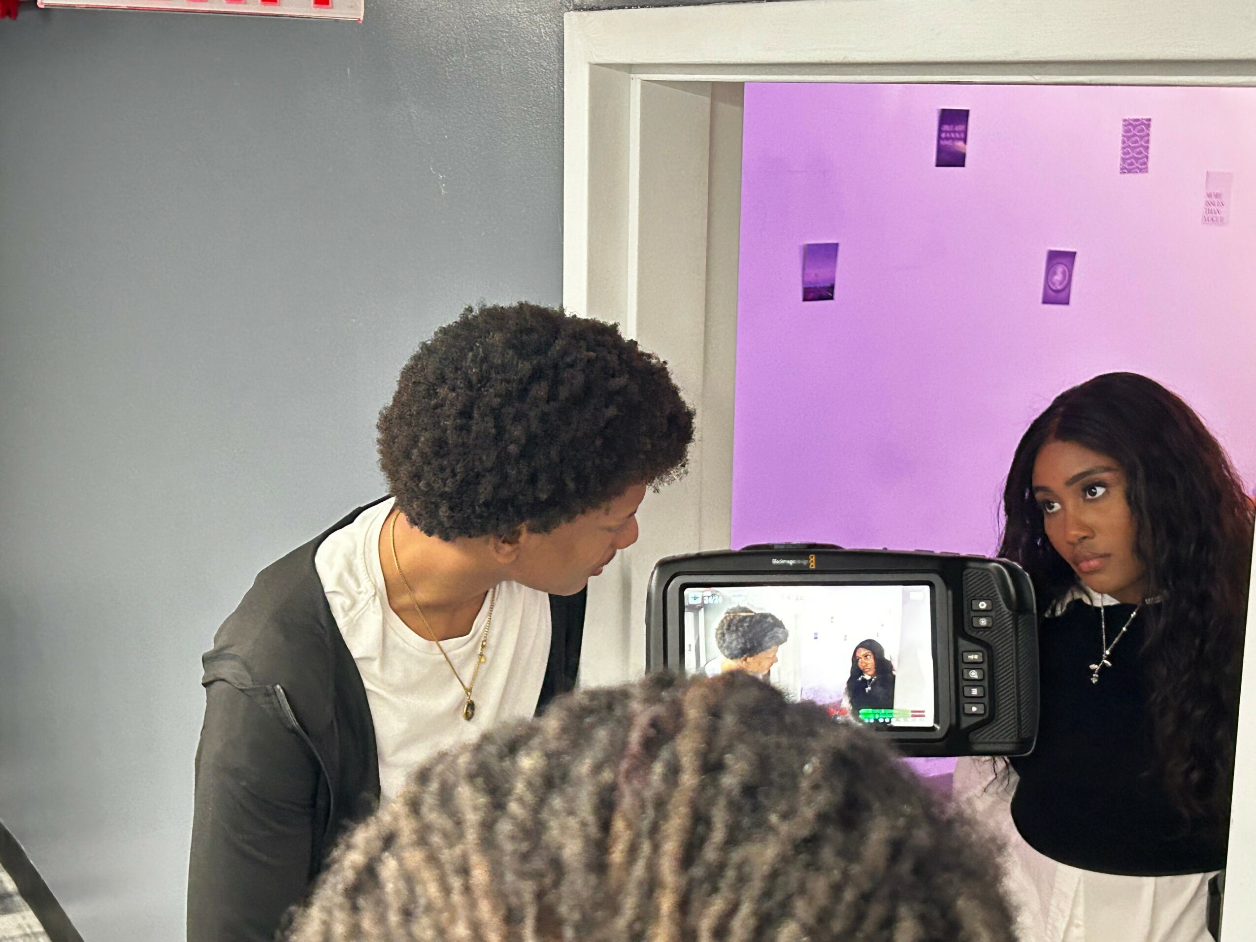 Two women are being recorded by a camera, one with curly hair and the other with straight hair, in a room with purple and gray walls.