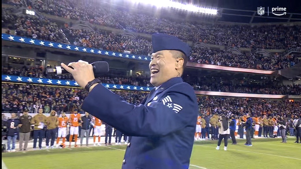 Person in uniform smiling and holding a microphone on a sports field with a crowd in the background.