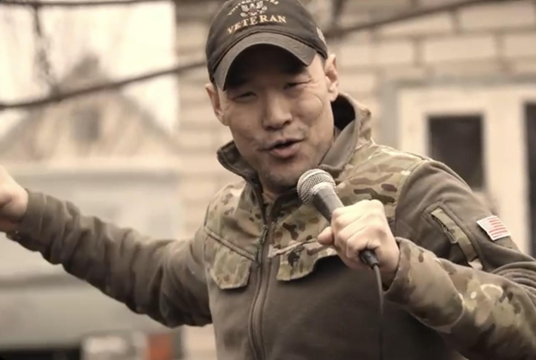 Person speaking into a microphone outdoors, wearing a camouflage jacket and cap, with a building in the background.