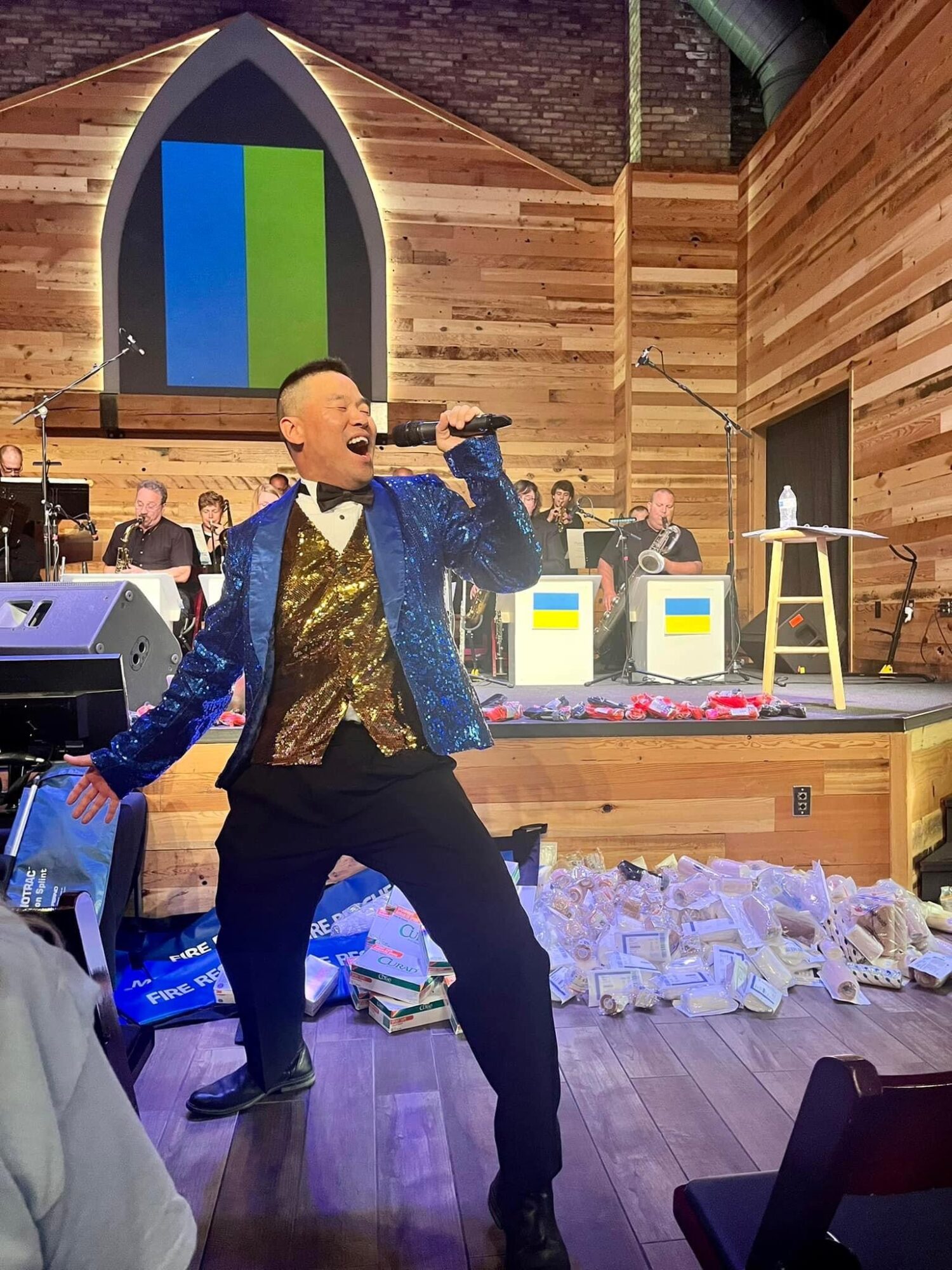 Performer singing passionately on stage with a microphone, band members in background, wooden interior, colorful decorations, and scattered items on floor.