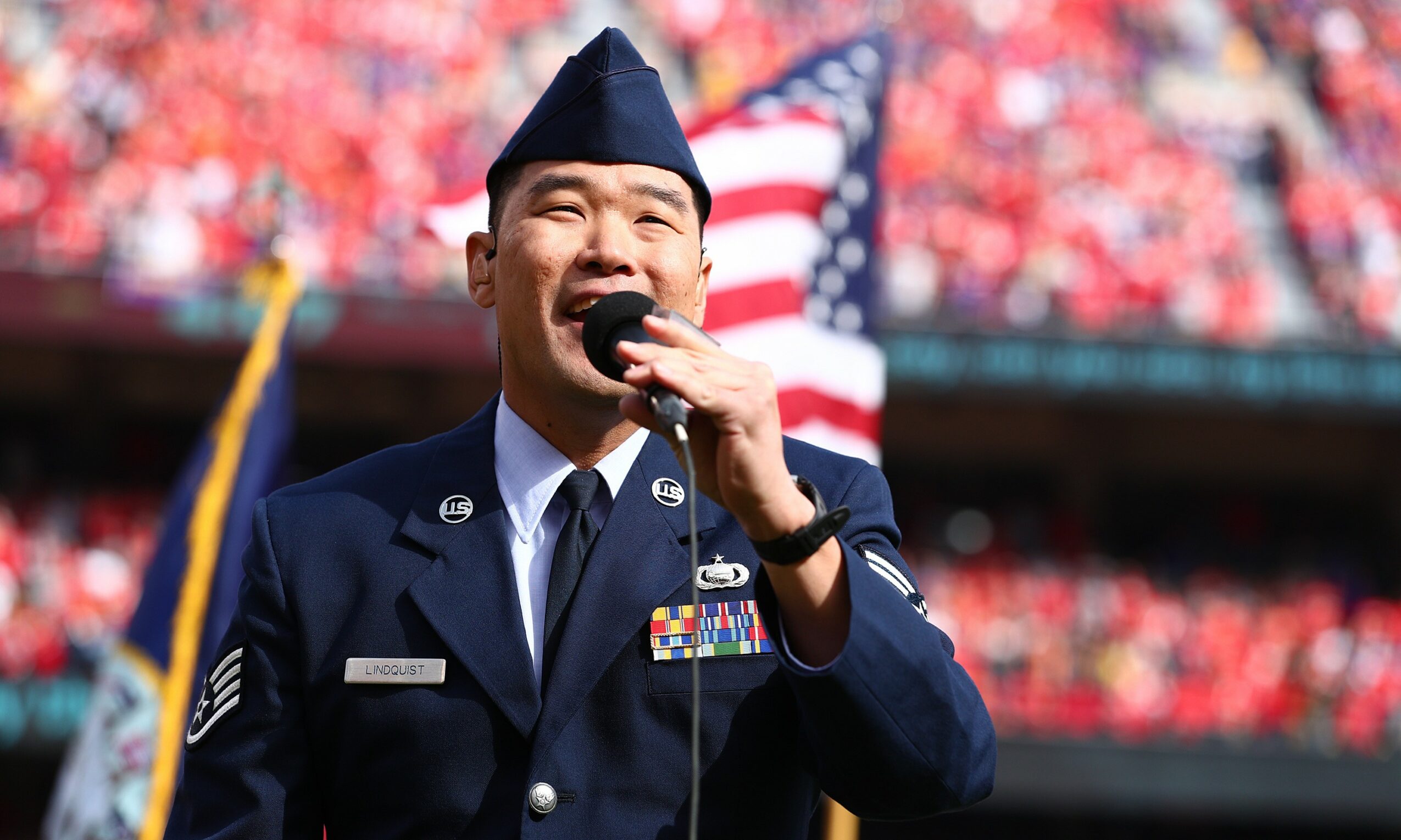A person in military uniform speaking into a microphone at a patriotic event, with a crowd and American flag in background.