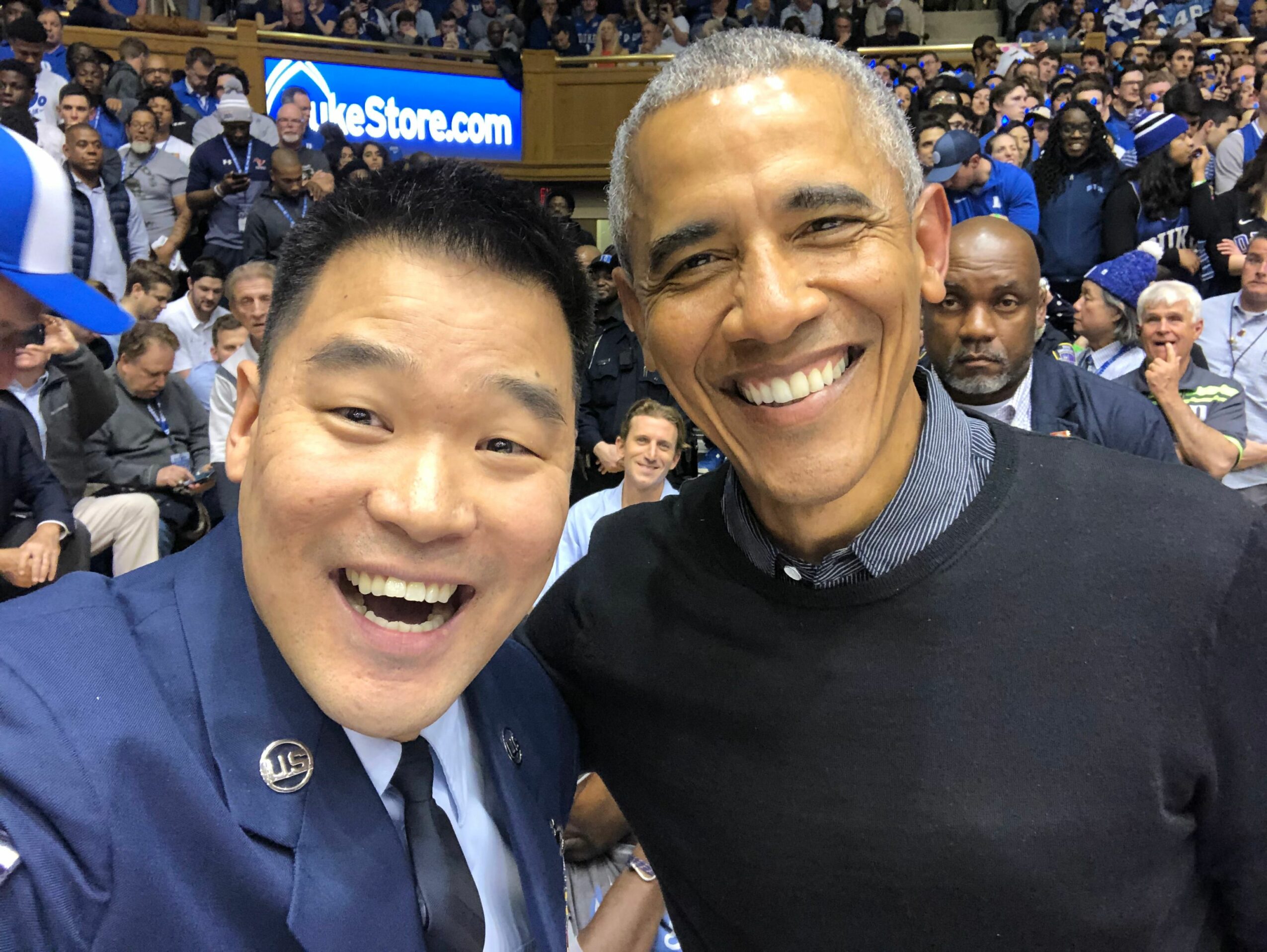 Two smiling people taking a selfie at a crowded outdoor event, with many people in the background.