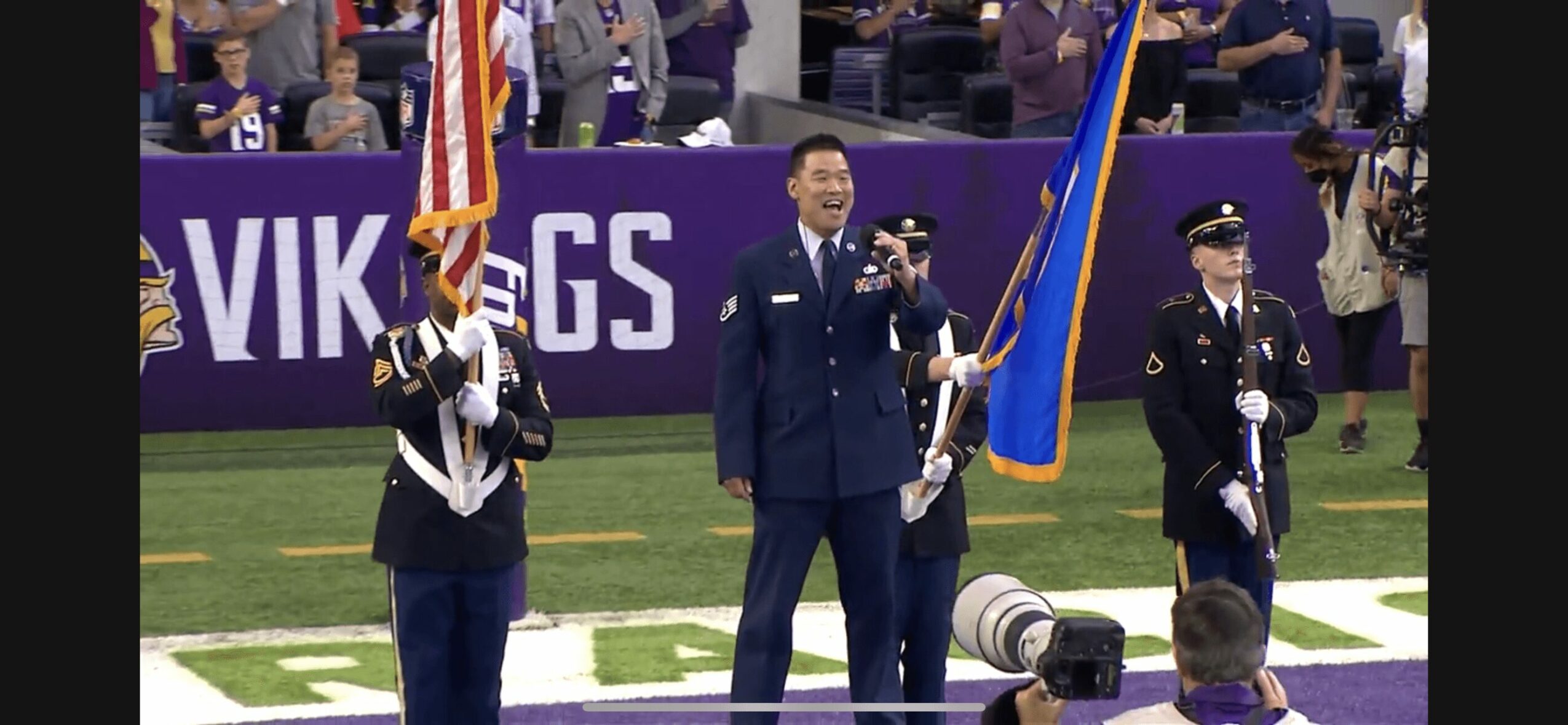 Three uniformed individuals holding flags on a field, with a person speaking into a microphone, and spectators in the background.