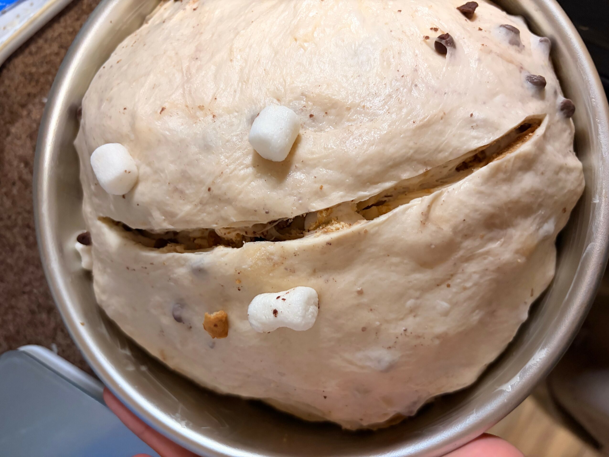 A round, smiling face-shaped bread with marshmallows for eyes and a mouth, in a metal bowl.