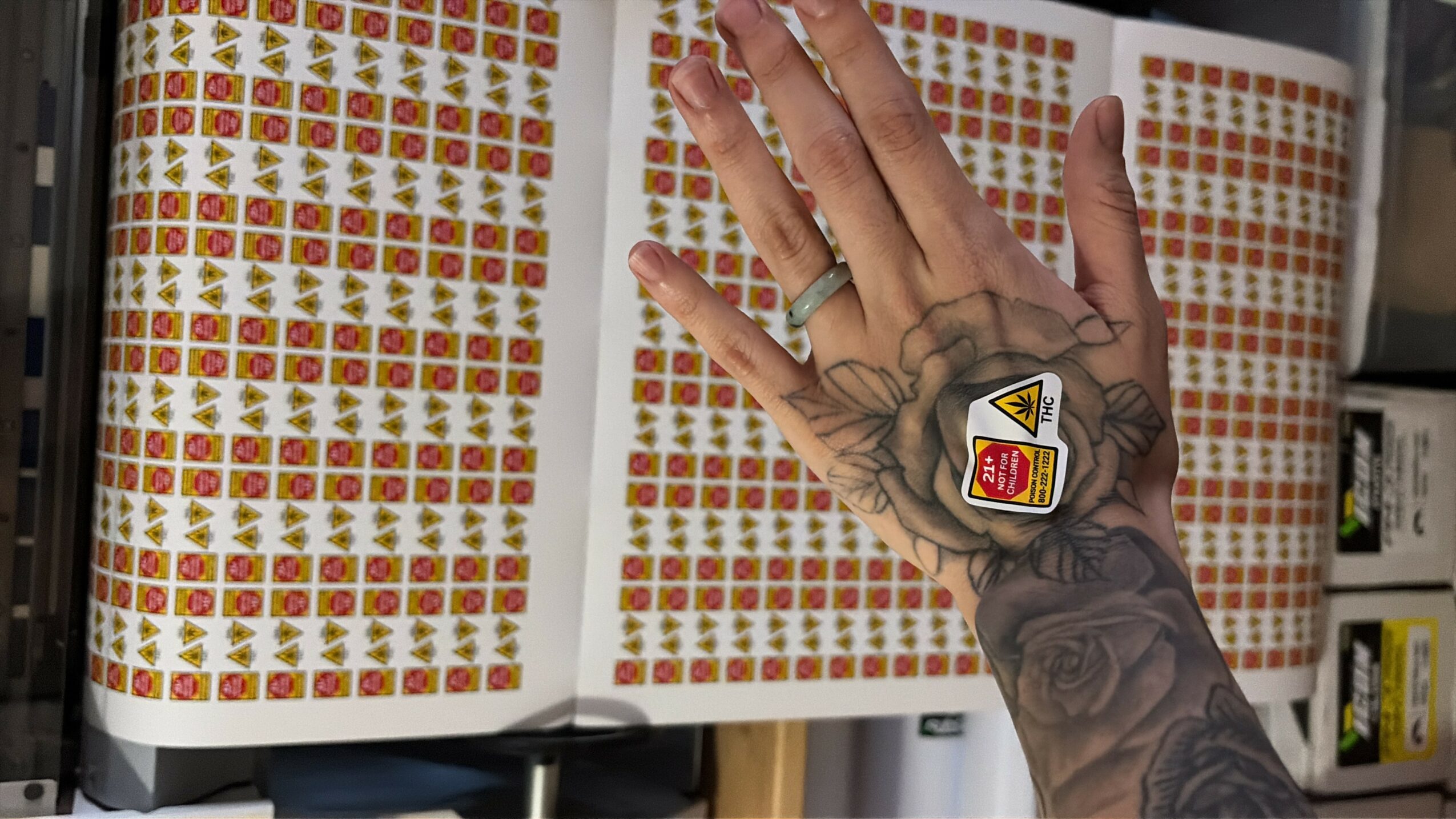 Hand with tattoo and ring holding open a book with colorful grid patterns, possibly stamps or stickers, on pages.