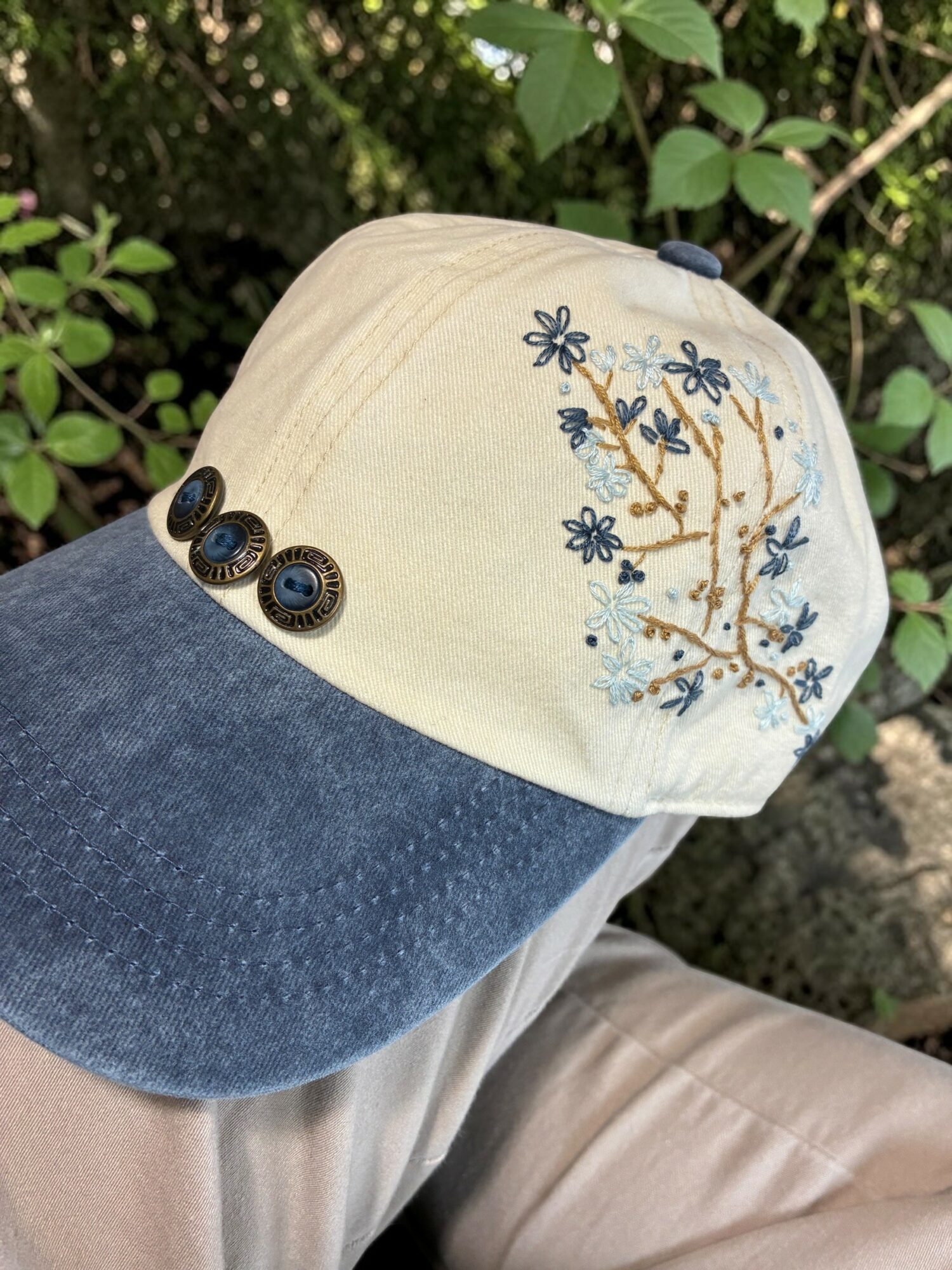 Cap with embroidered flowers and branches, beige top, dark blue brim, buttons on side, outdoors with green leaves