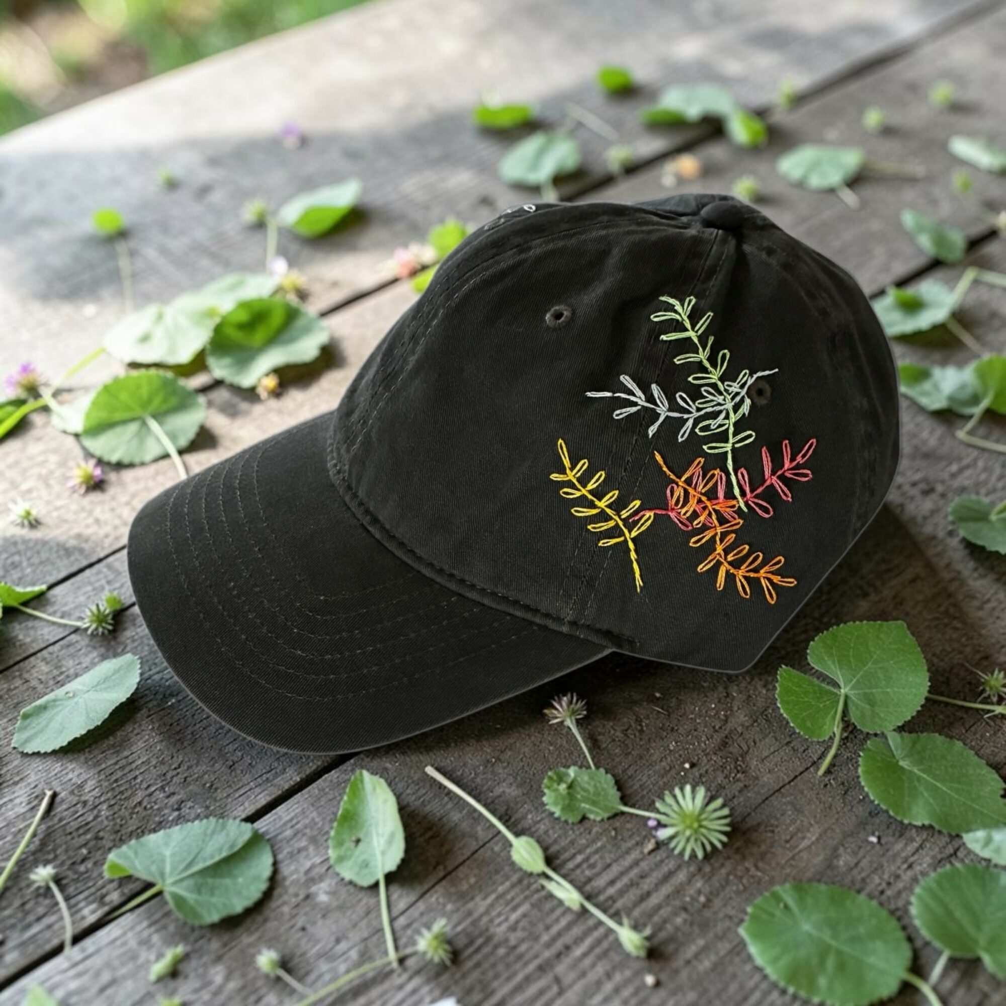 Black cap with colorful embroidered leaf design resting on a wooden surface surrounded by green leaves and small flowers.