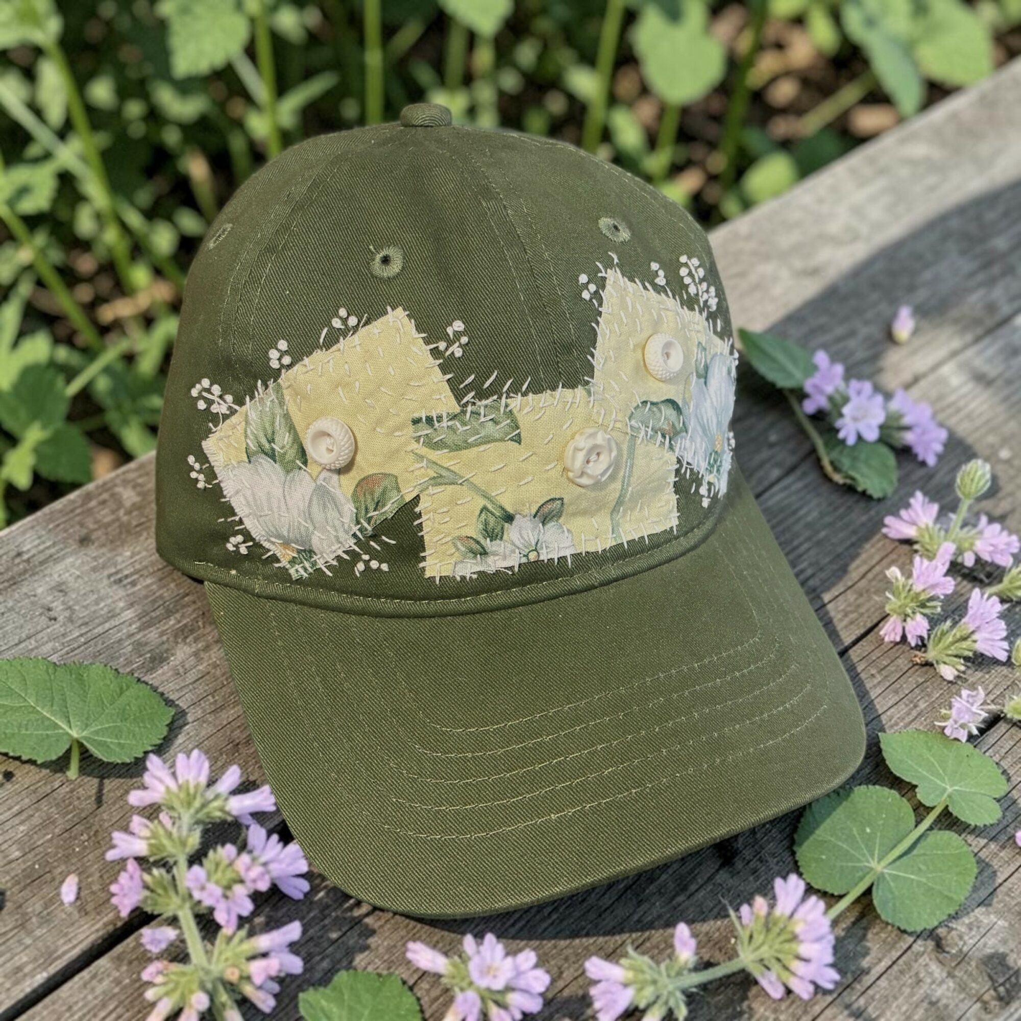 Olive green baseball cap with a nature-themed embroidered design, surrounded by purple flowers and green leaves on a wooden surface.