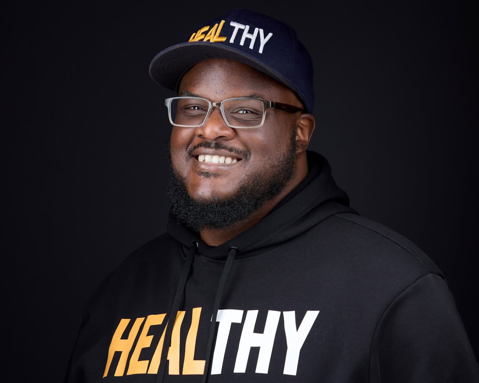 Smiling man wearing glasses, a cap, and a hoodie with the word 'HEALTHY' on it, against a dark background.