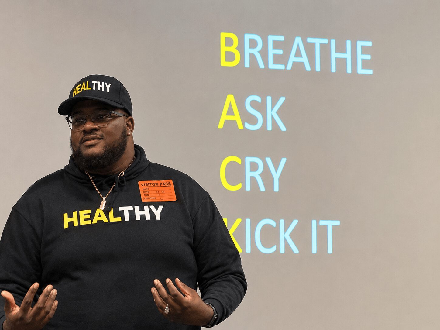 Man wearing a black hoodie and cap with 'HEALTHY' text, standing in front of a screen displaying health tips.