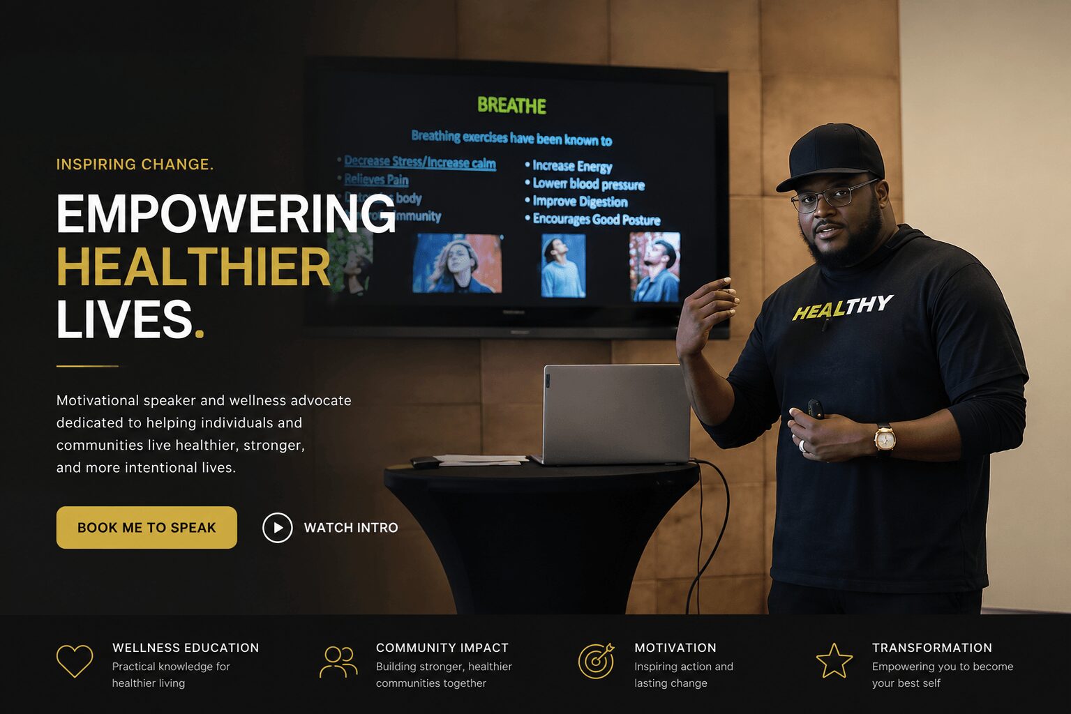 Man wearing a black shirt with 'Healthy' written on it, standing next to a laptop and presentation screen, in a wellness event setting.