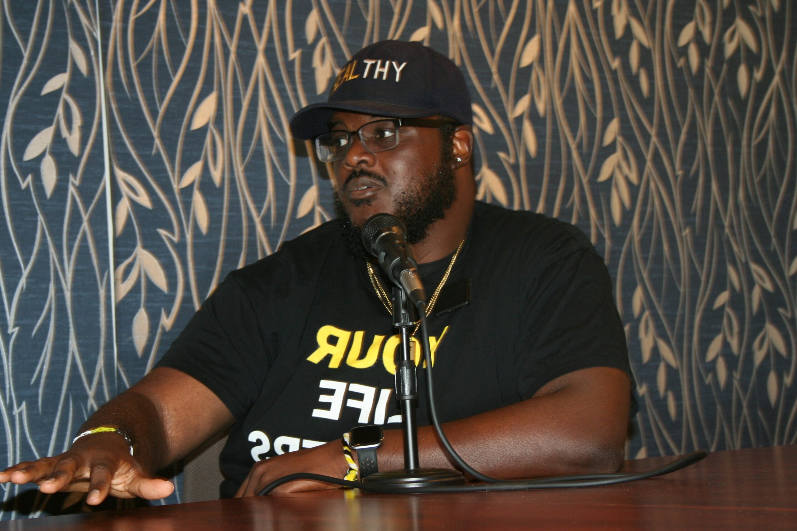 Person wearing a black cap and shirt, speaking into a microphone at a table, with patterned background.