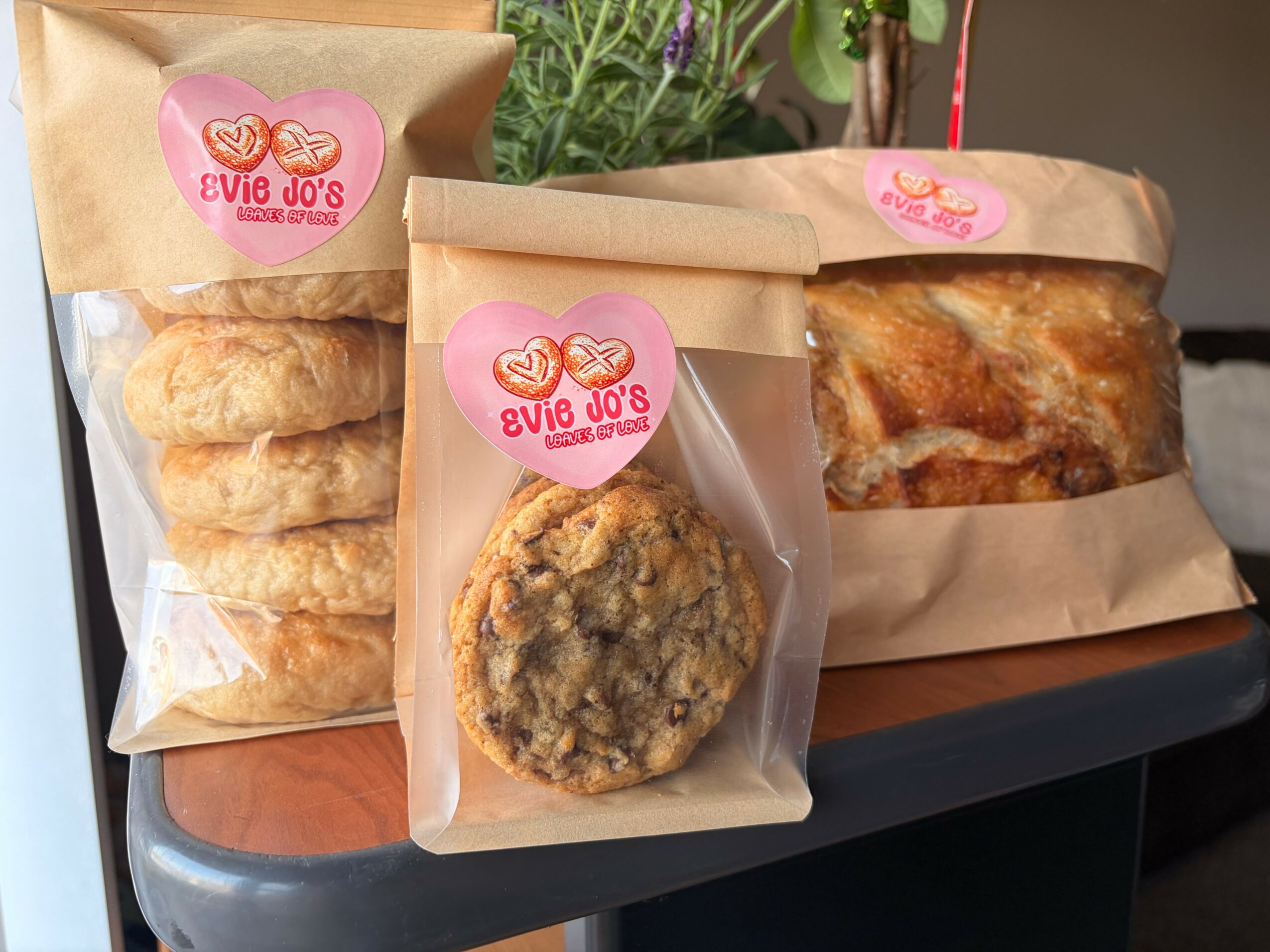 Three baked goods in paper bags with pink heart-shaped labels, one cookie in a clear bag, and a loaf in a paper bag, on a dark surface.