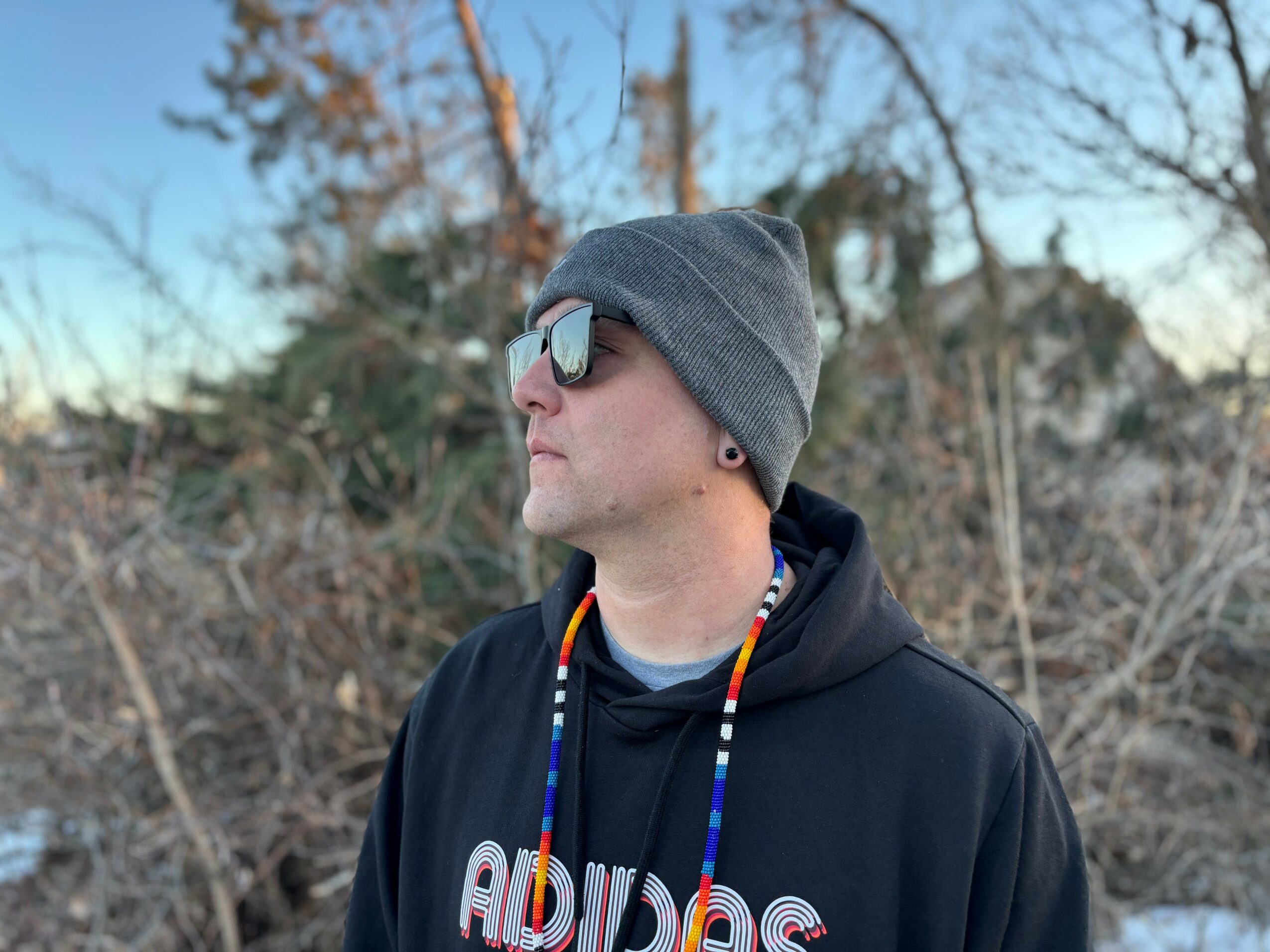 Person wearing sunglasses, gray beanie, and black hoodie outdoors with trees in background.