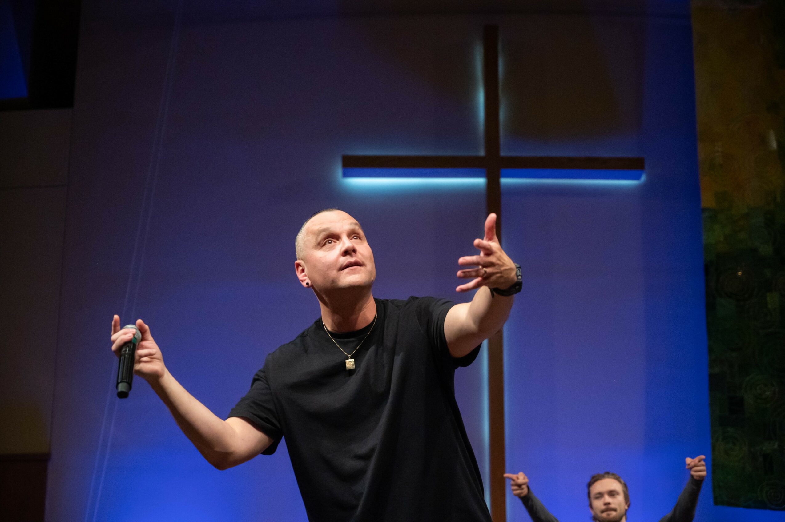 Person on stage holding microphone, gesturing upward, with illuminated cross in background, blue lighting, and another person partially visible.
