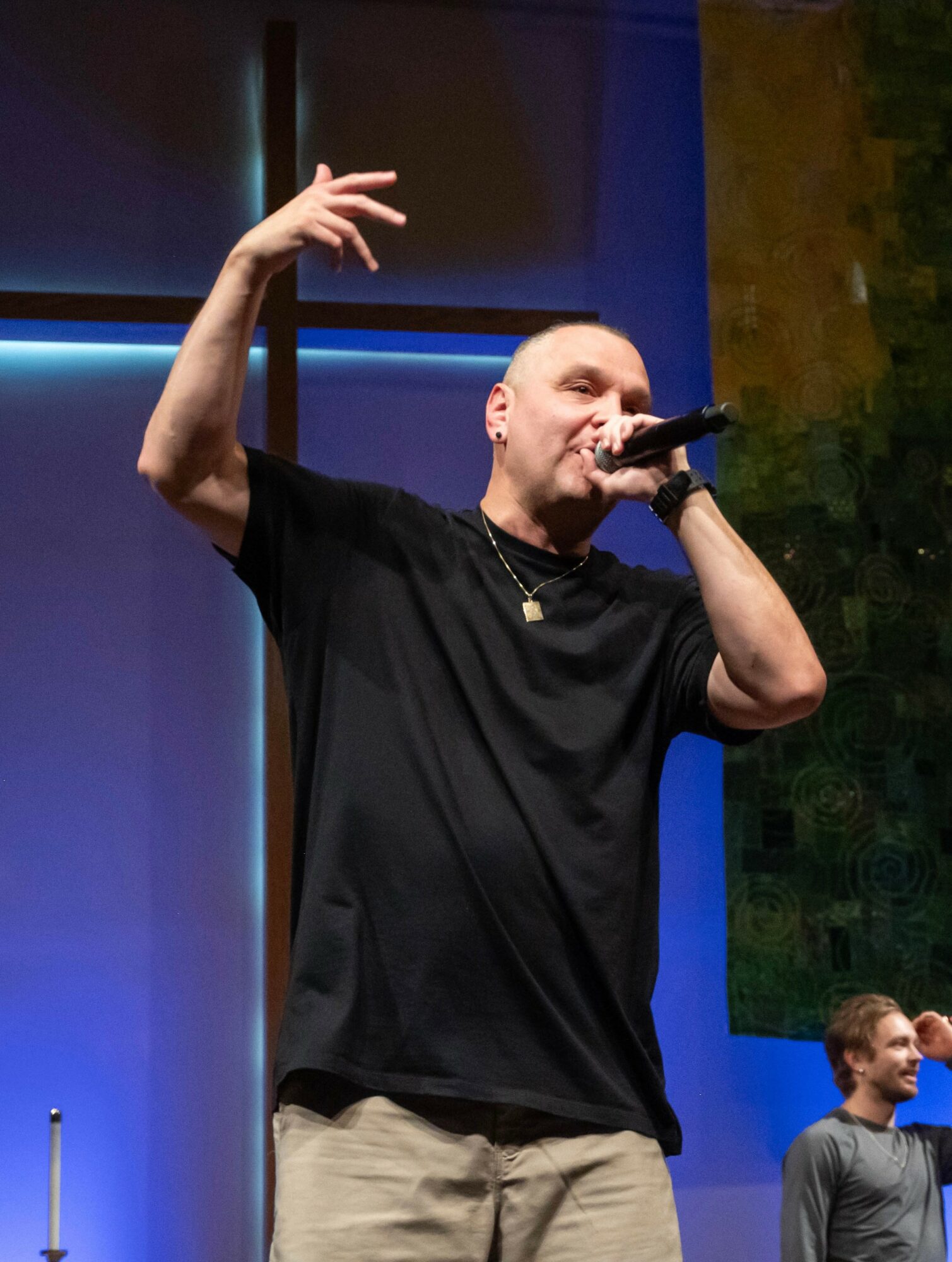 Male performer holding microphone, gesturing with hand, on stage with colorful background, wearing black shirt and beige pants.