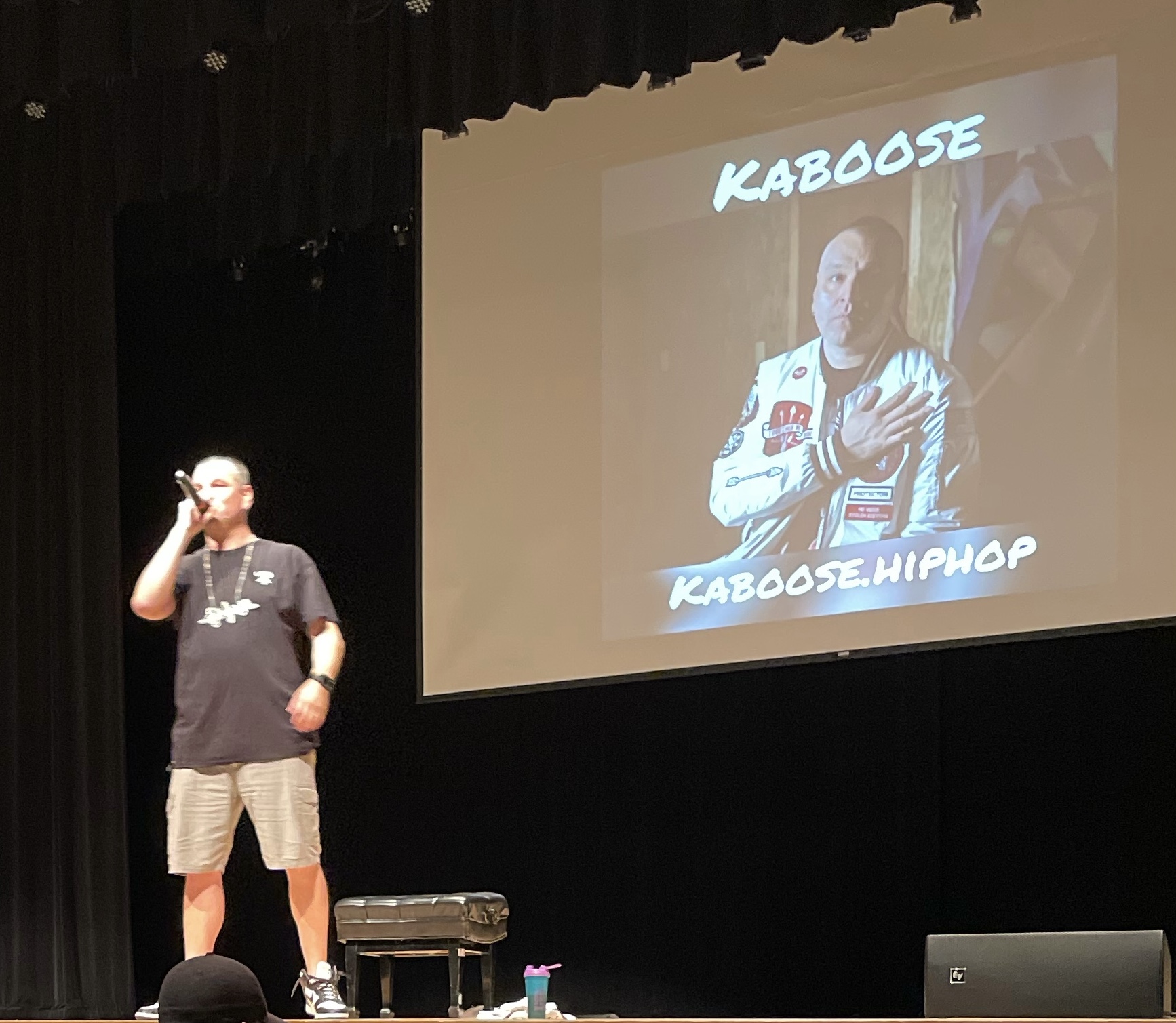 Person standing on stage speaking into microphone, large screen behind displaying a person in a jacket with text 'KABOOSE' and 'KABOOSE HIPHOP'.