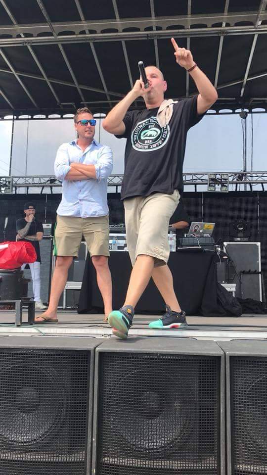 Two men on stage, one speaking into a microphone, the other standing with arms crossed, in an outdoor setting.