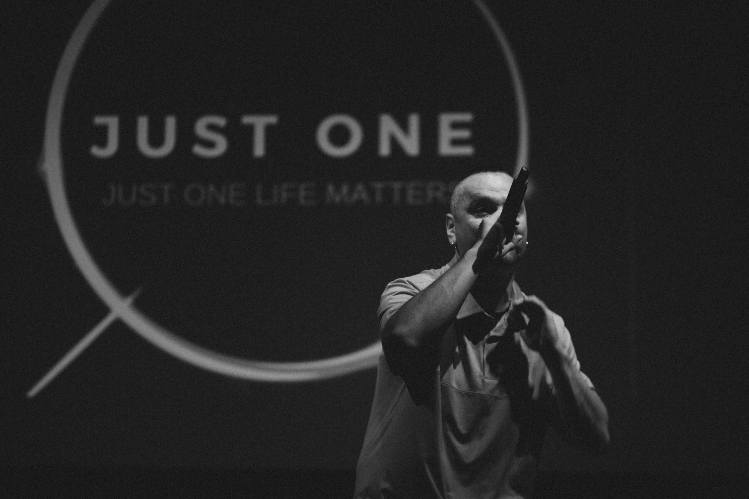 Person holding a microphone in front of a screen displaying the text 'JUST ONE' and 'JUST ONE LIFE MATTERS'.