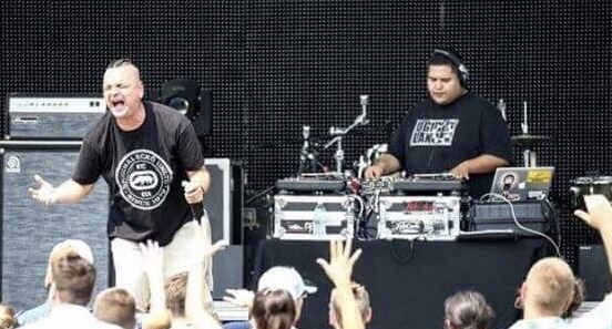 Two performers on stage, one singing and the other DJing, with an audience in front, some raising hands.