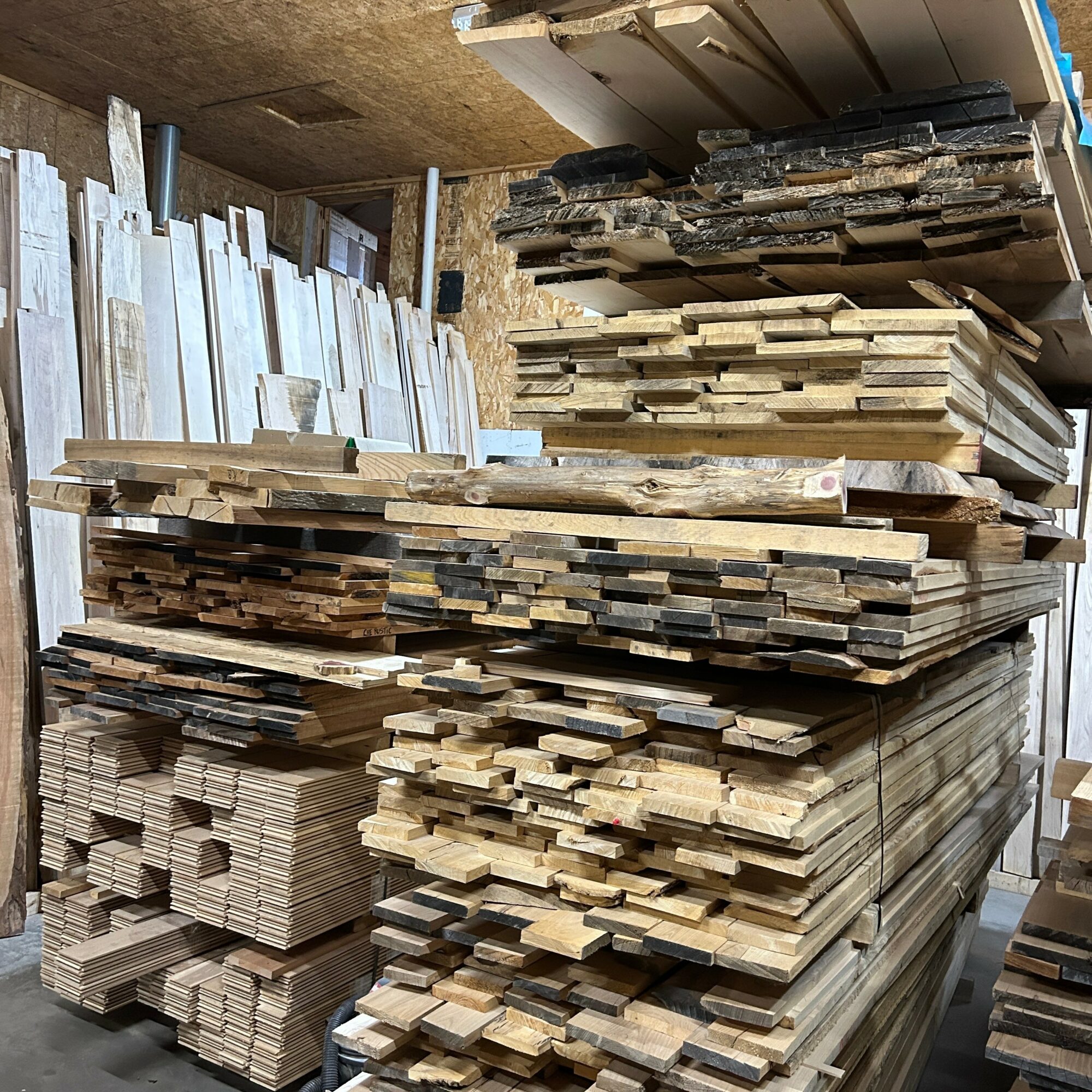 Stacks of wooden planks and boards in a workshop or storage area, organized in piles on shelves and the floor.