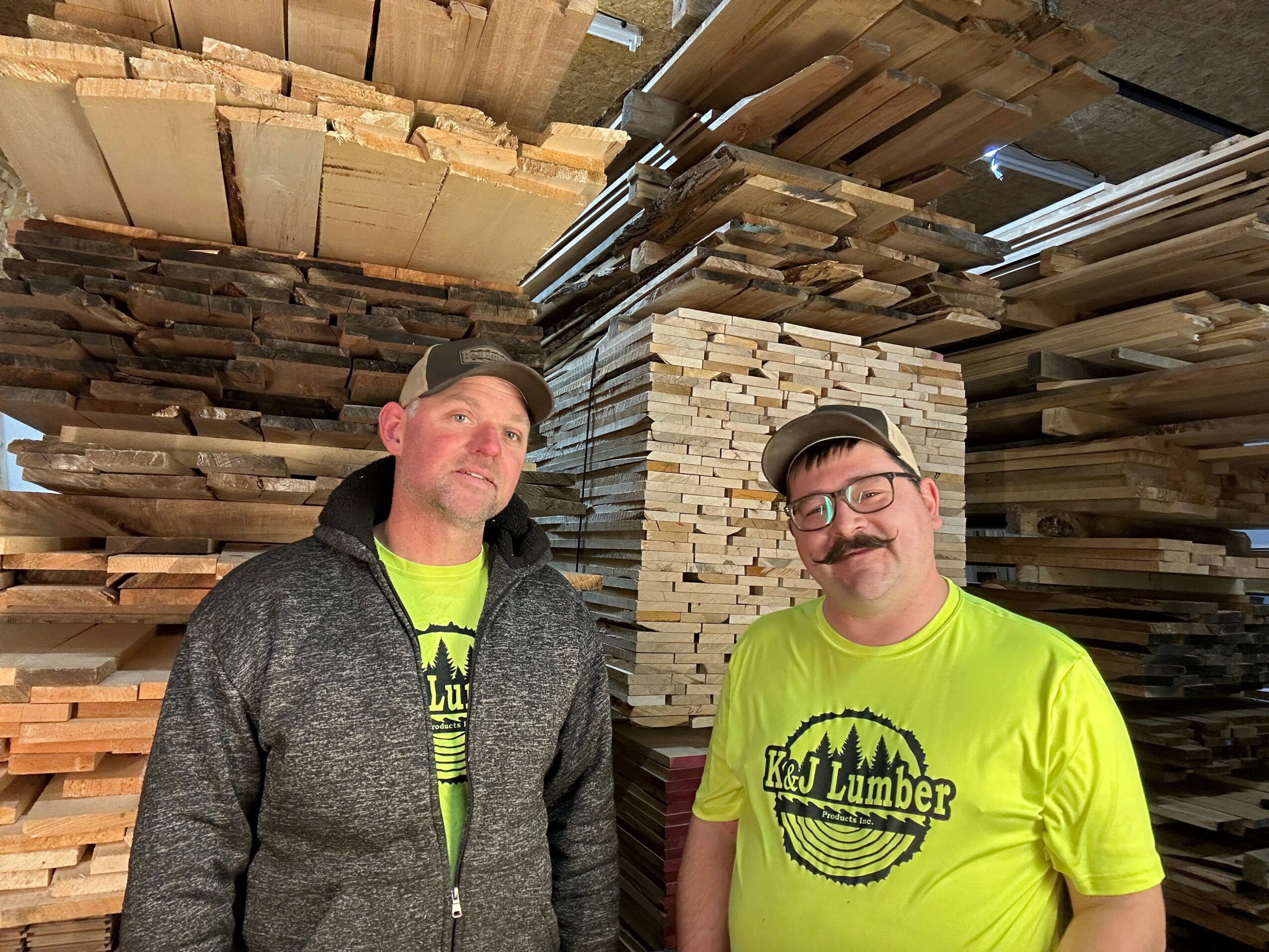 Two men in bright yellow shirts stand in front of stacked lumber in a warehouse.