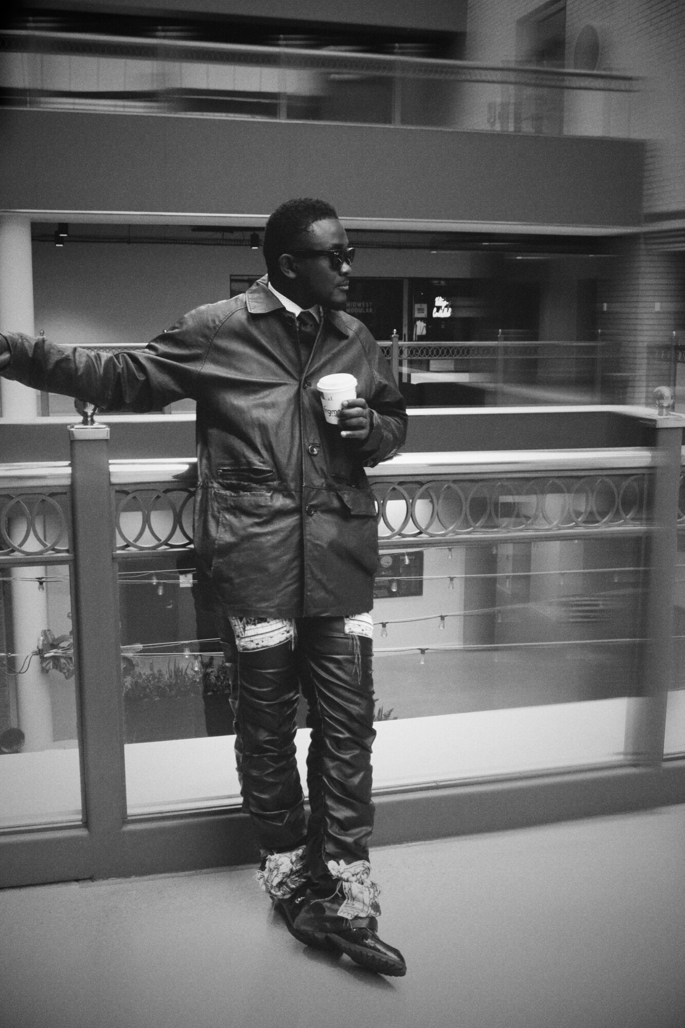 Person standing on a balcony railing, holding a cup, in an indoor setting with multiple levels visible behind.