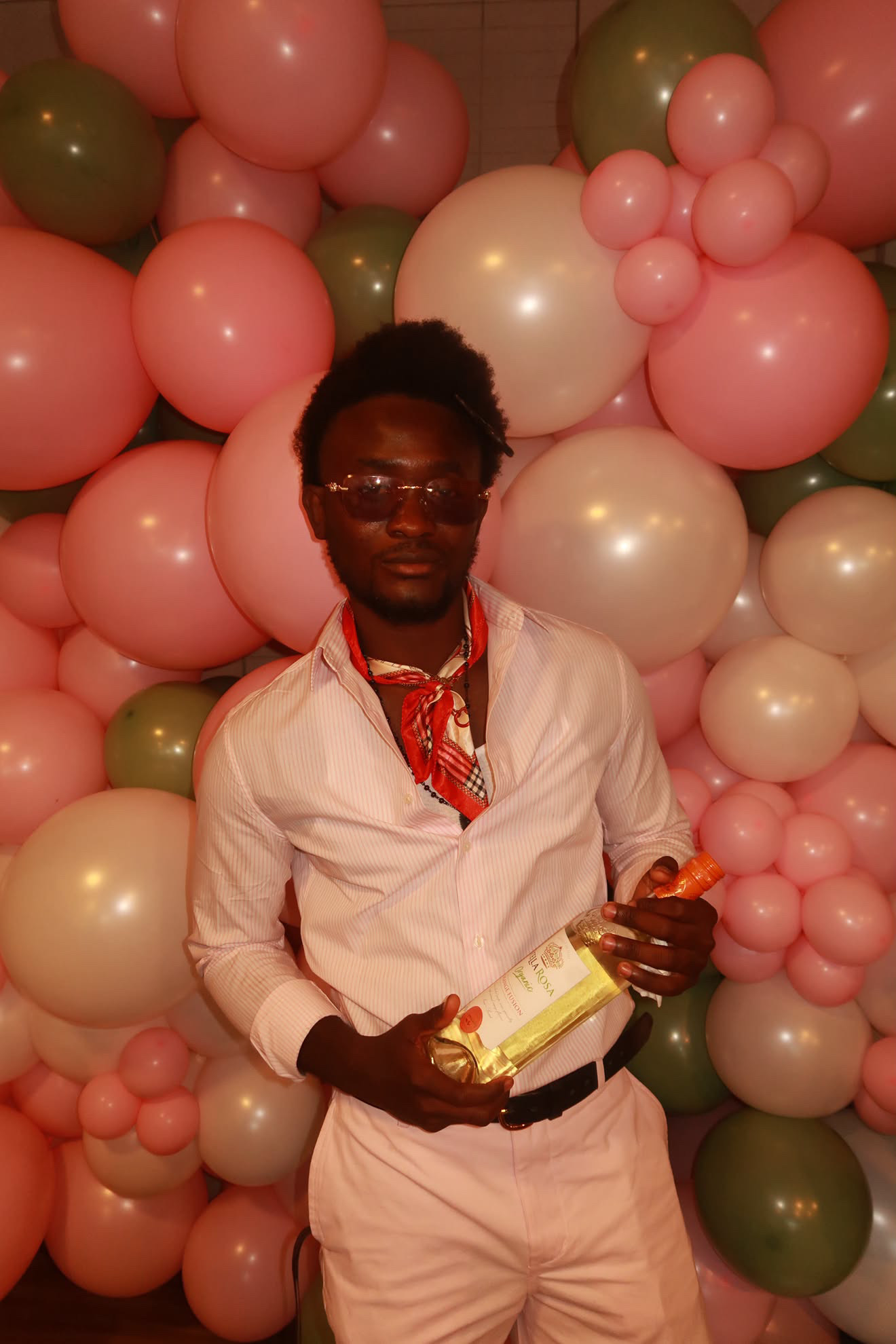 Person with dark skin and glasses holding a gift, standing in front of pink, white, and green balloons.