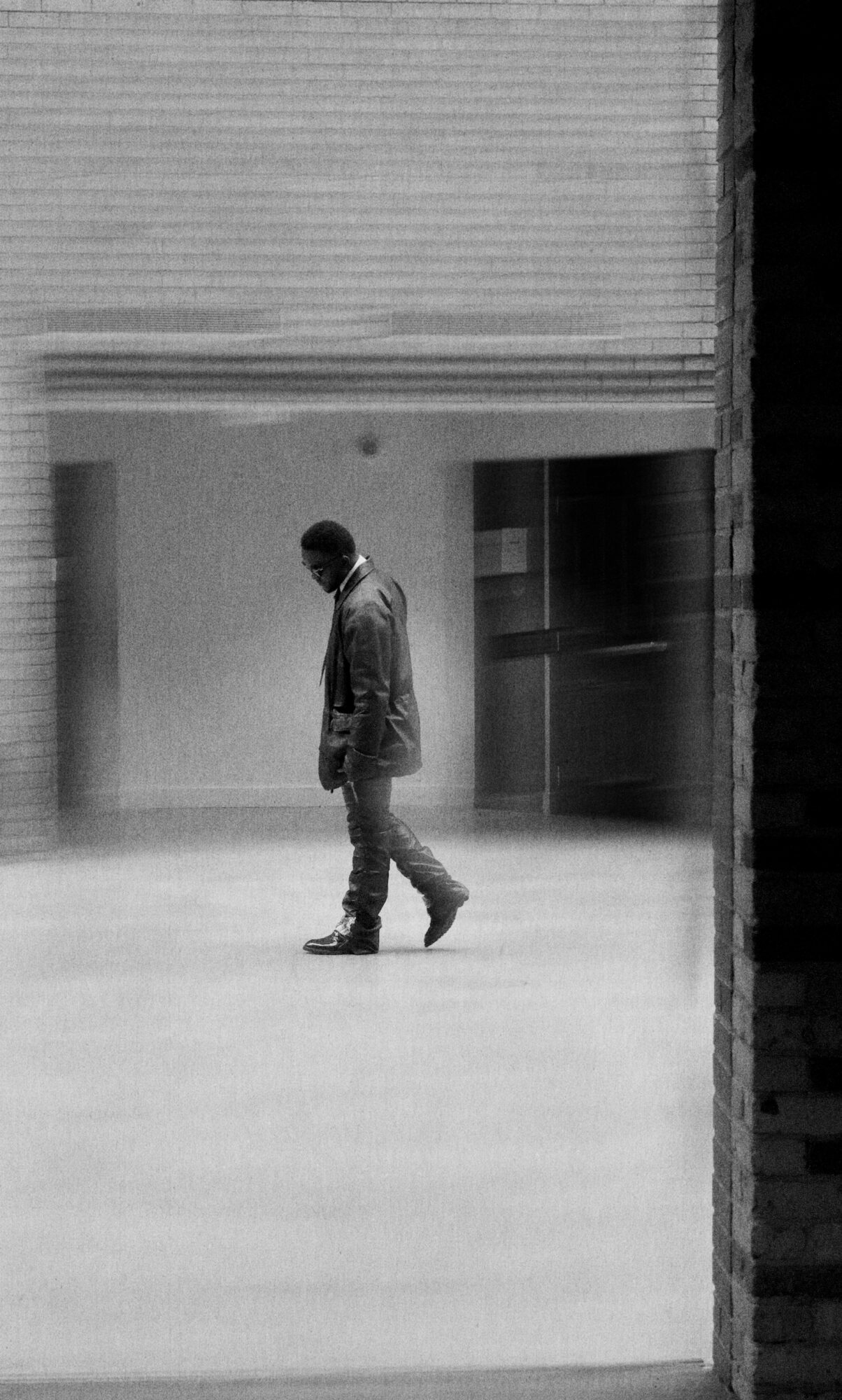 A person walking indoors, seen through a window or opening, with a modern building background.