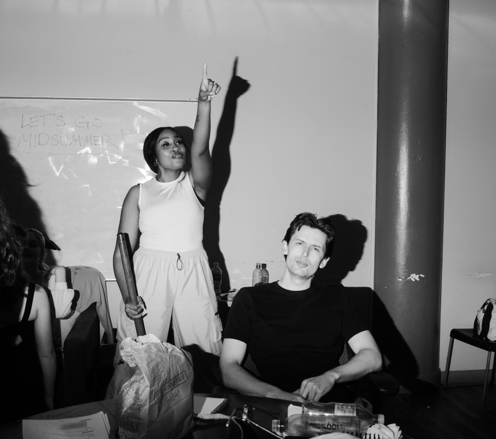 A woman stands with her arm raised, pointing upward, while a man sits at a table with a neutral expression.