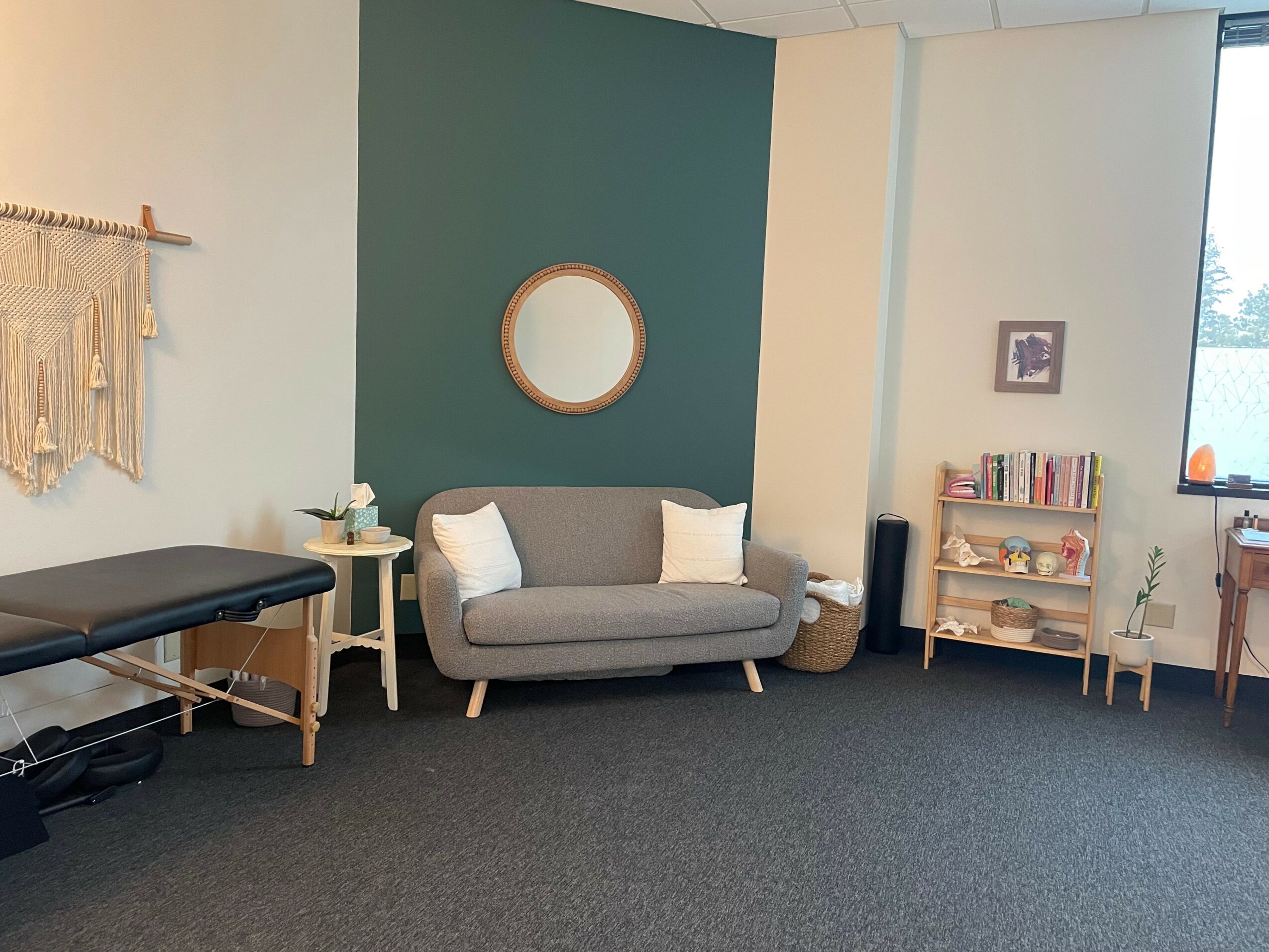 Room with a gray sofa, round mirror, bookshelf, window, and a massage table, with a dark carpeted floor.