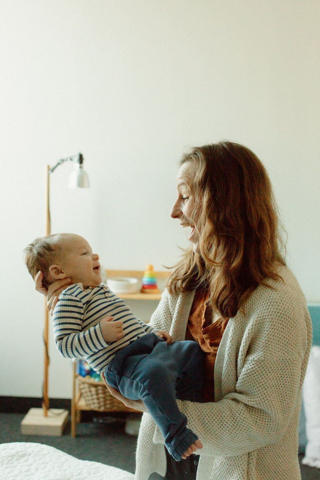 Woman holding a baby in a room with a lamp and basket, smiling at each other, light-colored wall background.
