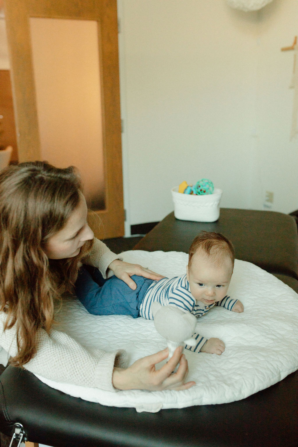 Woman with long hair lying on bed with baby on stomach, baby wearing striped shirt, in a room with a basket of toys in the background.