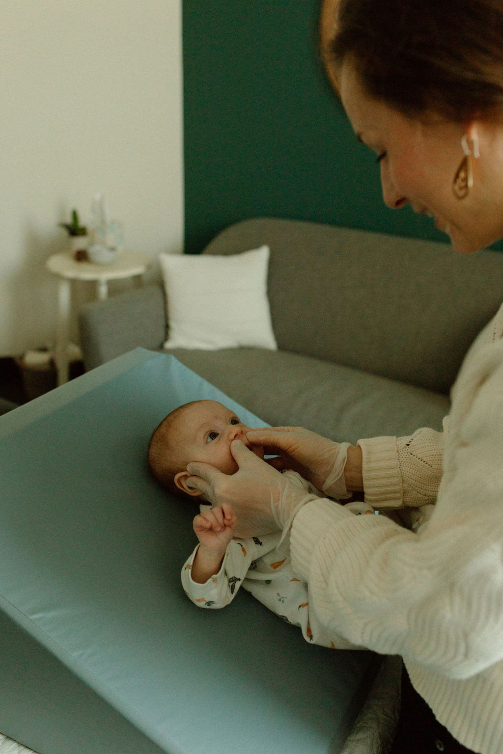 Woman gently holds a baby on a changing table in a living room, with a sofa and side table in the background.