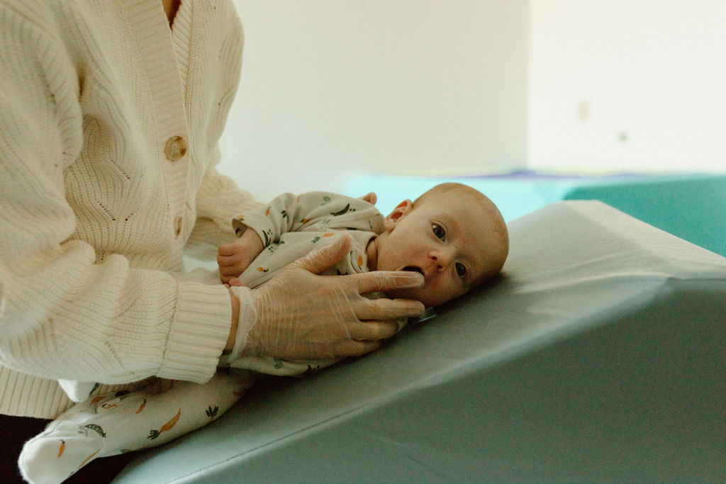 A baby lying on a hospital bed with a person gently holding the baby's head, in a bright room.