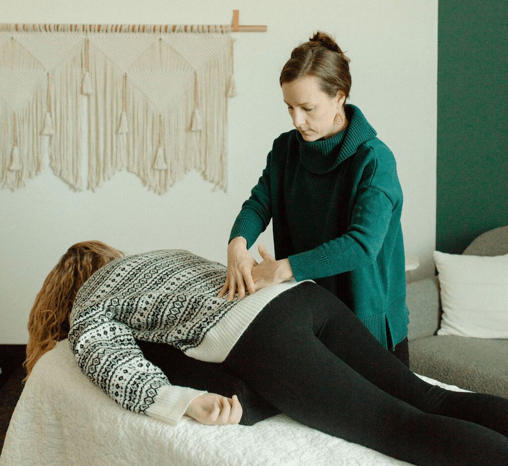 Woman receiving massage from another woman in a room with wall hanging and sofa, lying on bed face down.