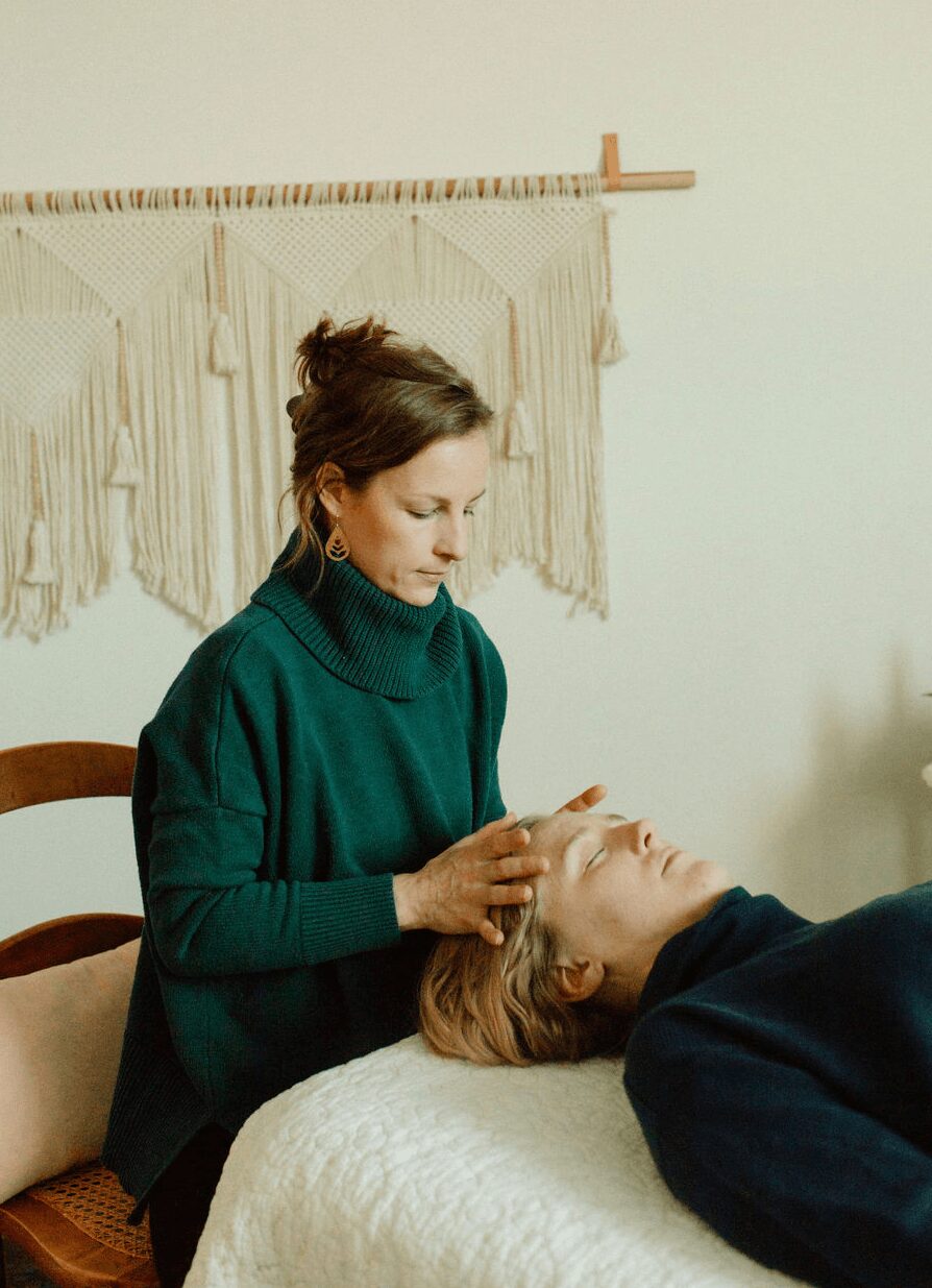 Woman with brown hair in a bun wearing a green sweater, performing a facial on a person lying down, in a room with a wall hanging.