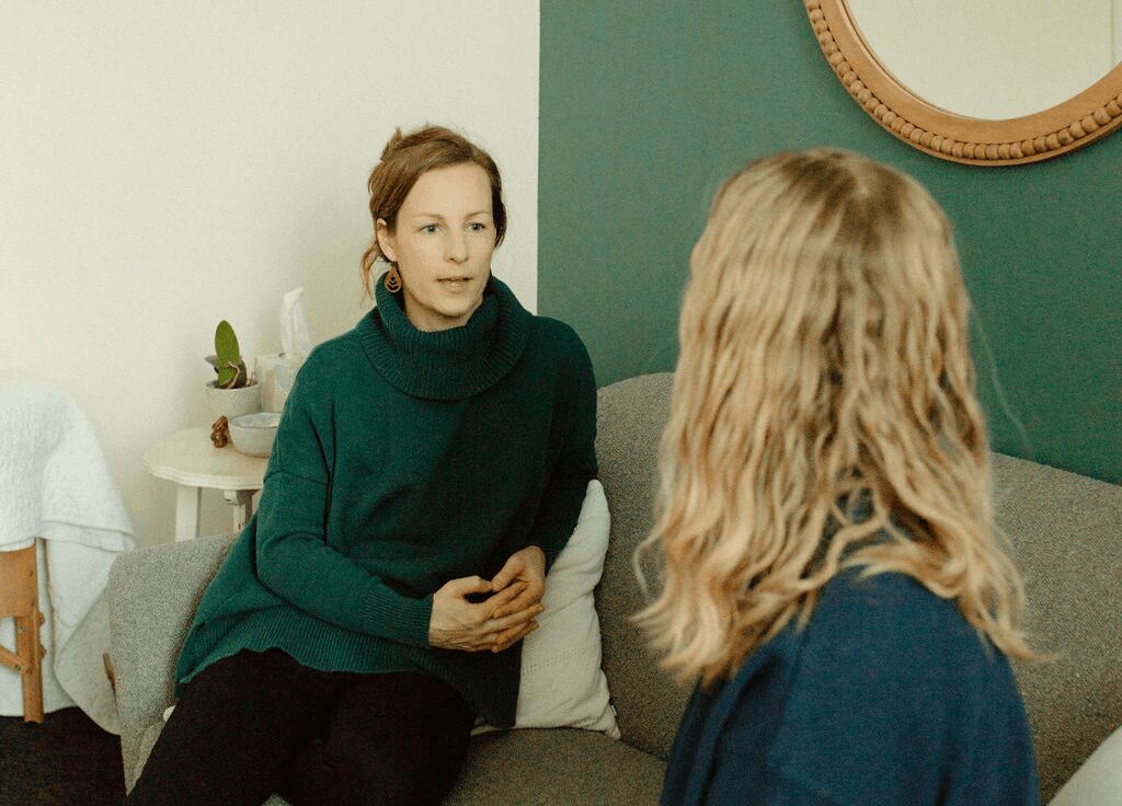 Two women sit on a sofa in a room with green and white walls, engaged in conversation.