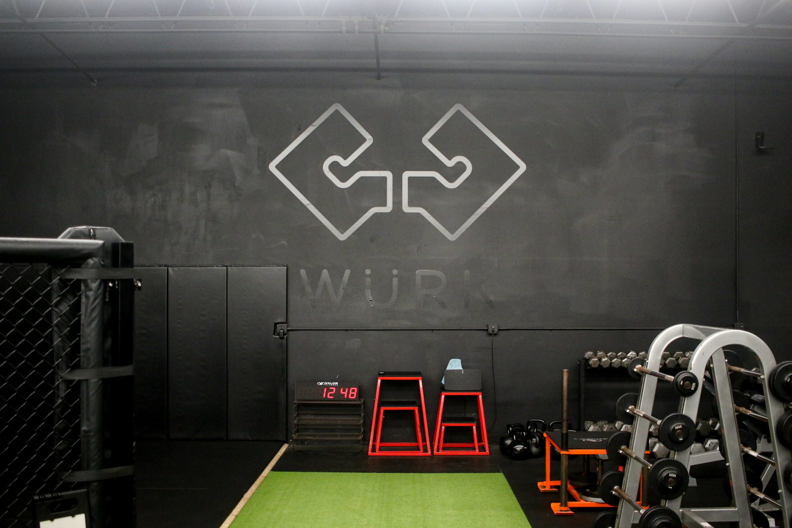 Gym with black wall, logo, and workout equipment, including a step platform and weights, on a green floor mat.