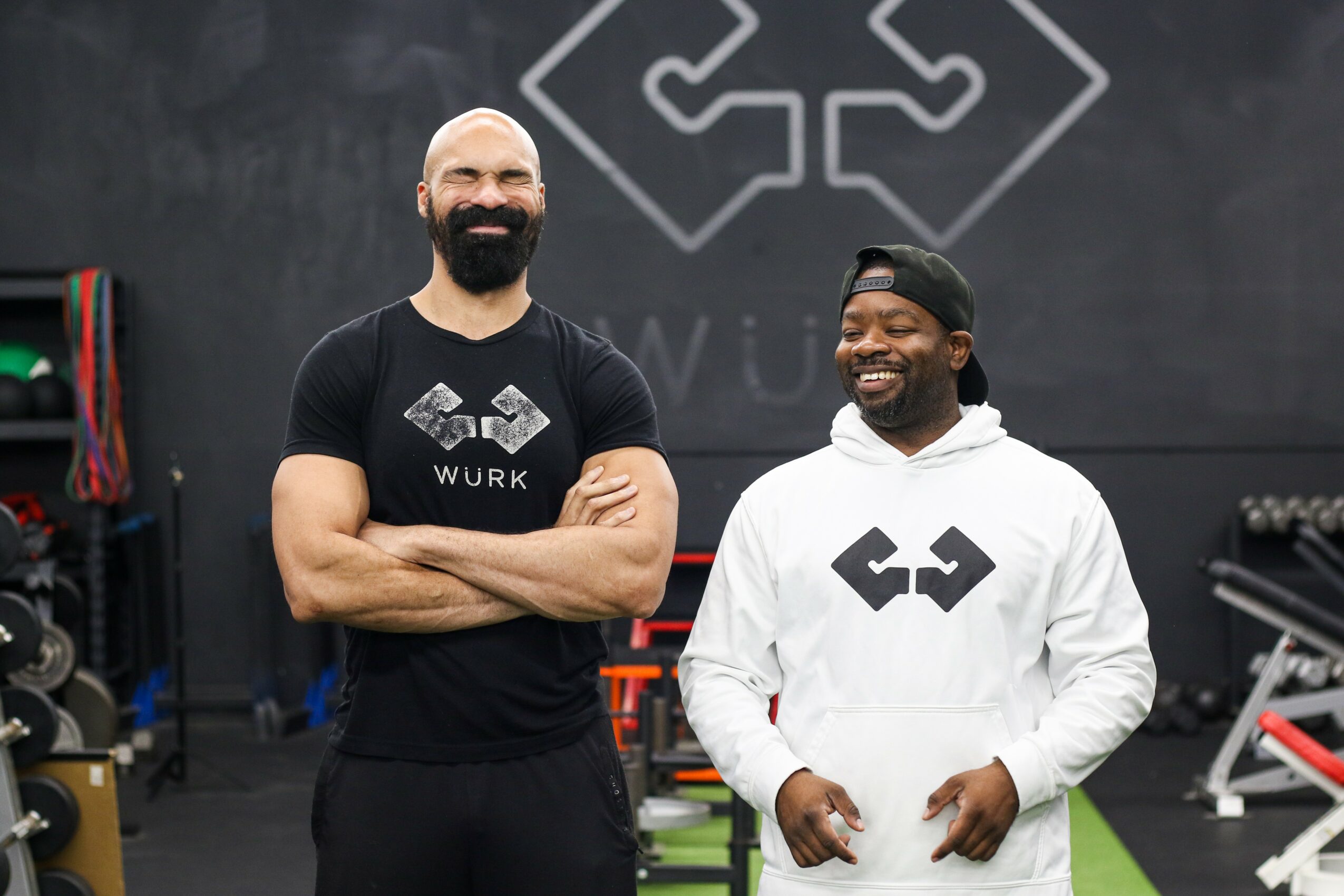 Two smiling men stand in a gym, one in a black shirt and the other in a white hoodie, with gym equipment in background.
