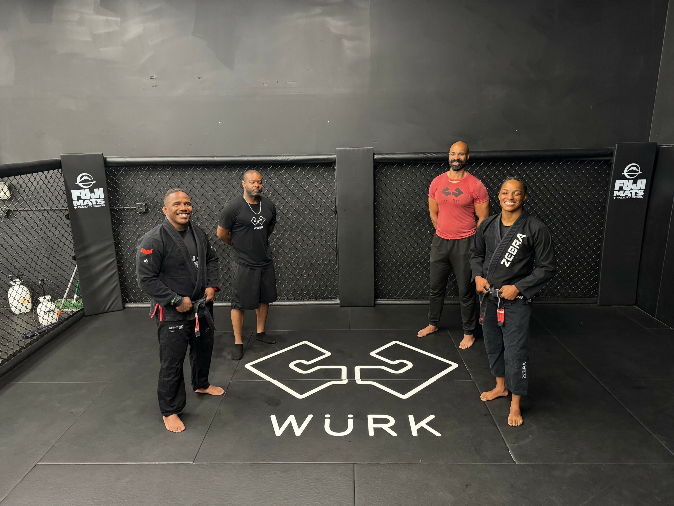 Four people in martial arts uniforms standing in a gym with a black wall and logo on the floor.