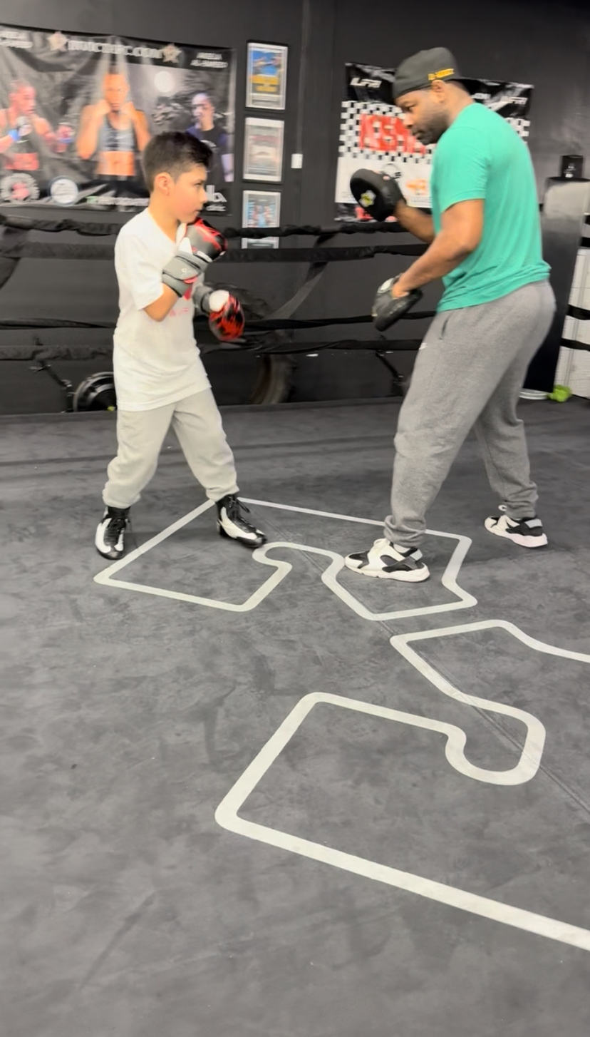 Young boy and adult sparring with boxing gloves in a gym, standing inside a white outlined boxing ring area.