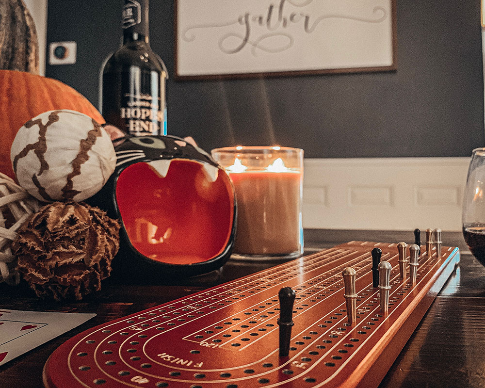 A shot of a wooden tabletop with a vintage roulette wheel, a candle, a bottle, and decorative pumpkins, with a wall and framed art in the background.
