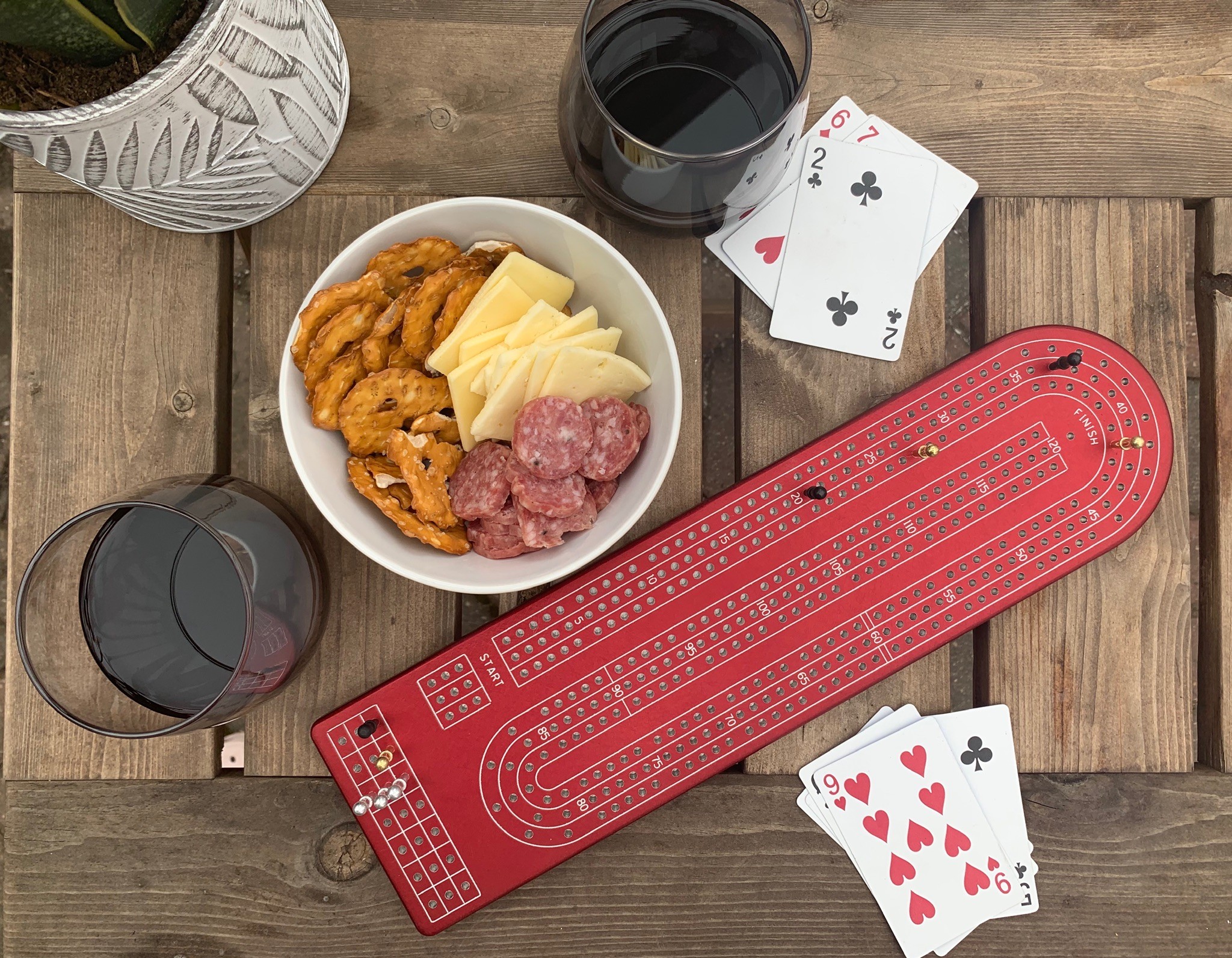 Table with a bowl of cheese, meat, and fried food, two glasses of red wine, a red gaming mat, and playing cards.