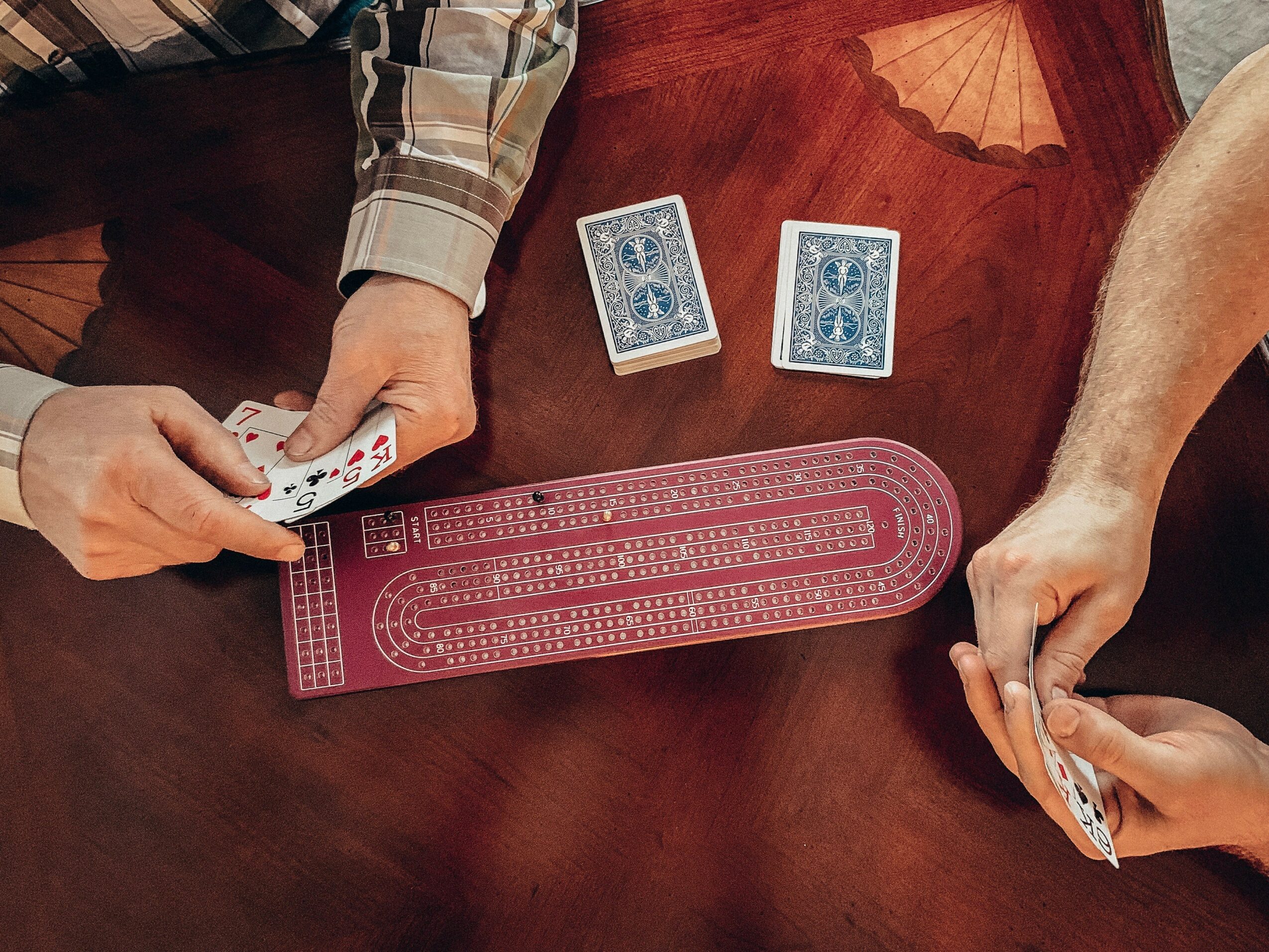 Two people play a card game at a table with a pink game board and two face-down cards, one person holding cards.