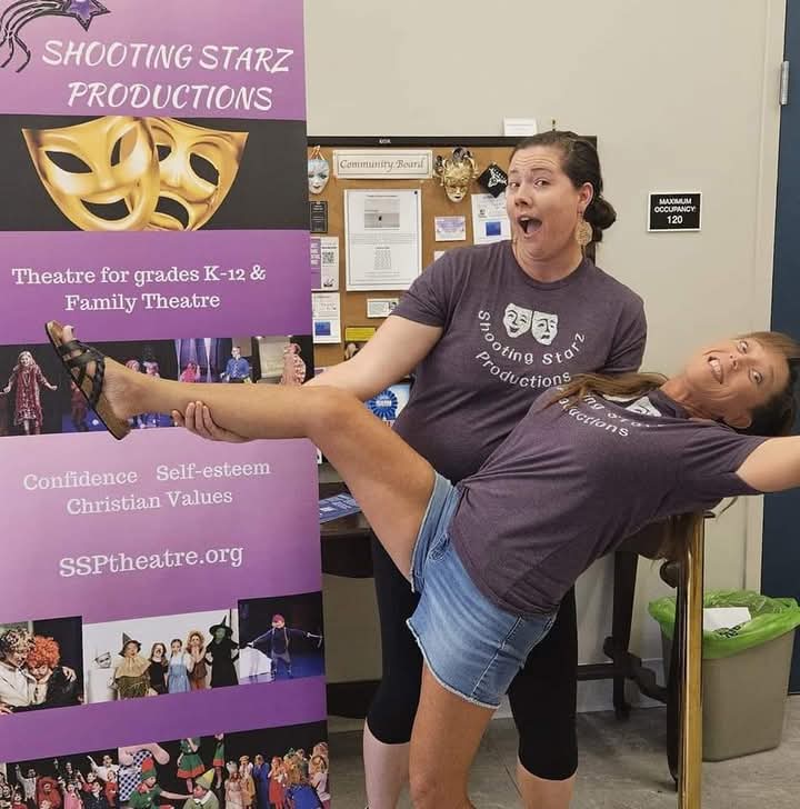 Two women in matching shirts, one holding the other's leg in a dance pose, in a room with a poster and bulletin board.